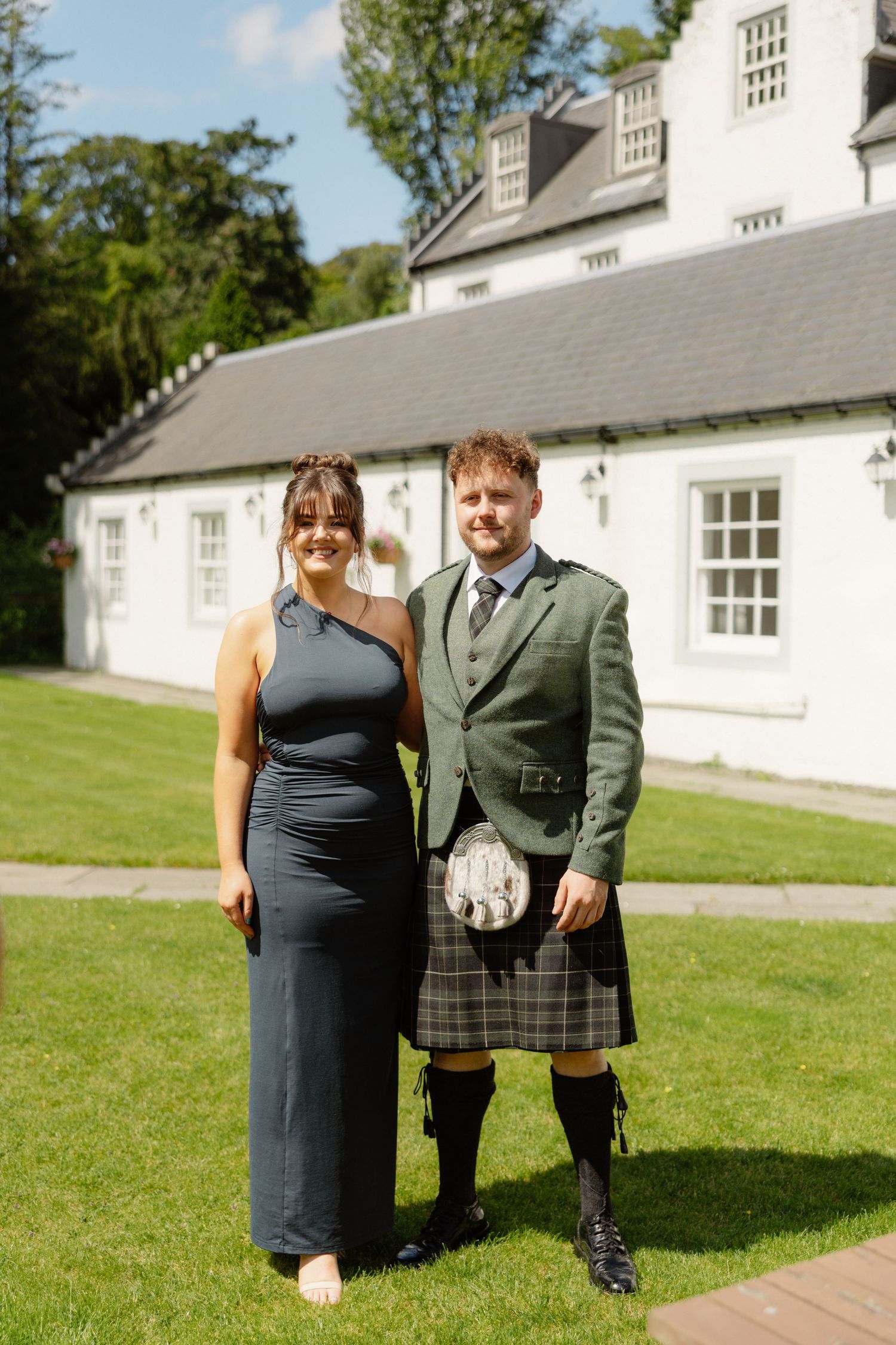 Woman in navy dress and man in traditional Scottish kilt and jacket posing outside white Scottish venue.