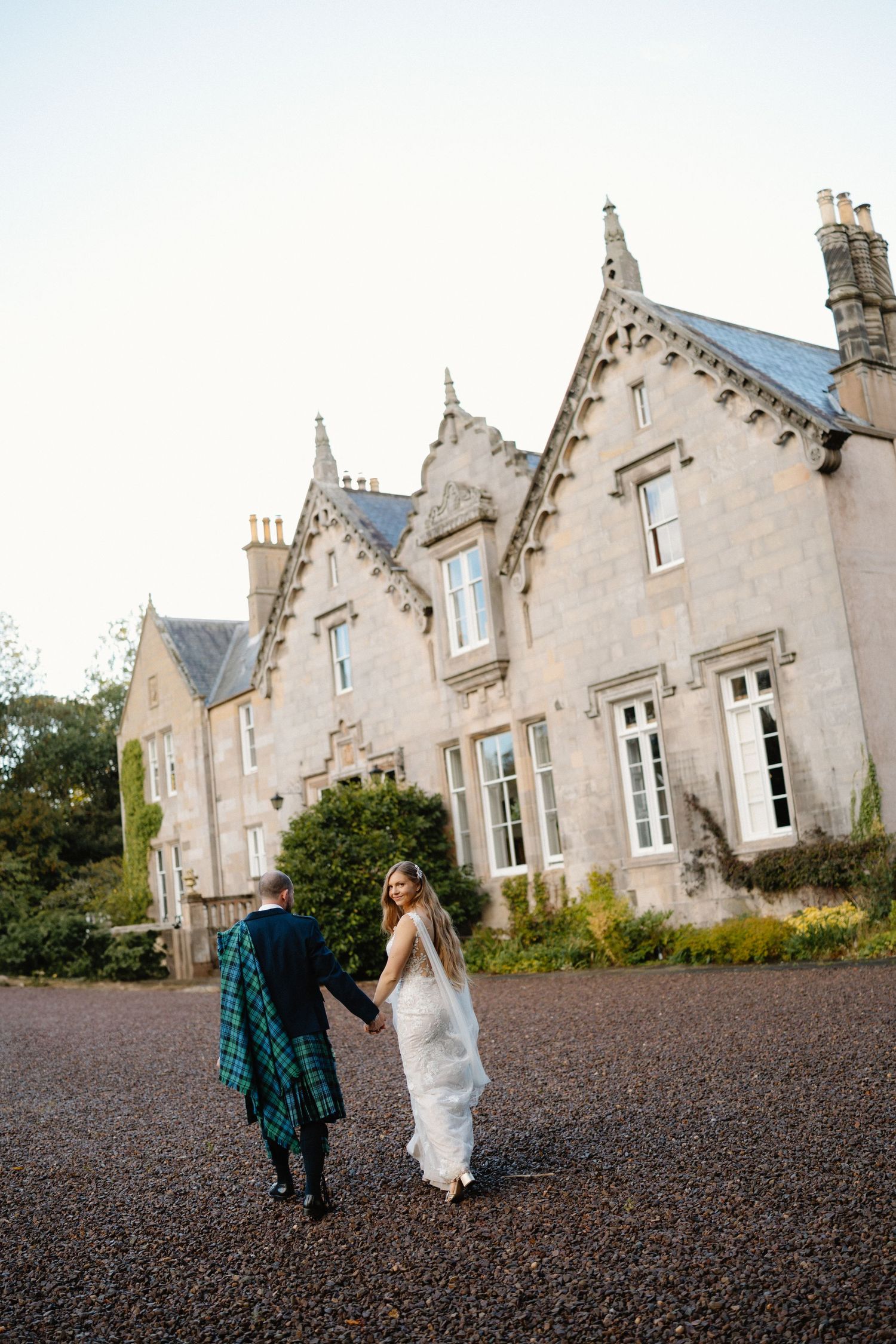 A couple walks hand in hand across the gravel courtyard of a grand Scottish Gothic-style manor house.