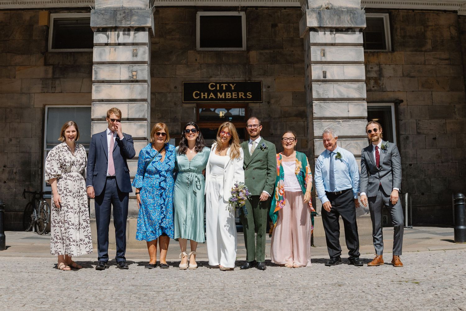Wedding party members lined up against stone wall in coordinated blue and neutral colored attire for group photo.