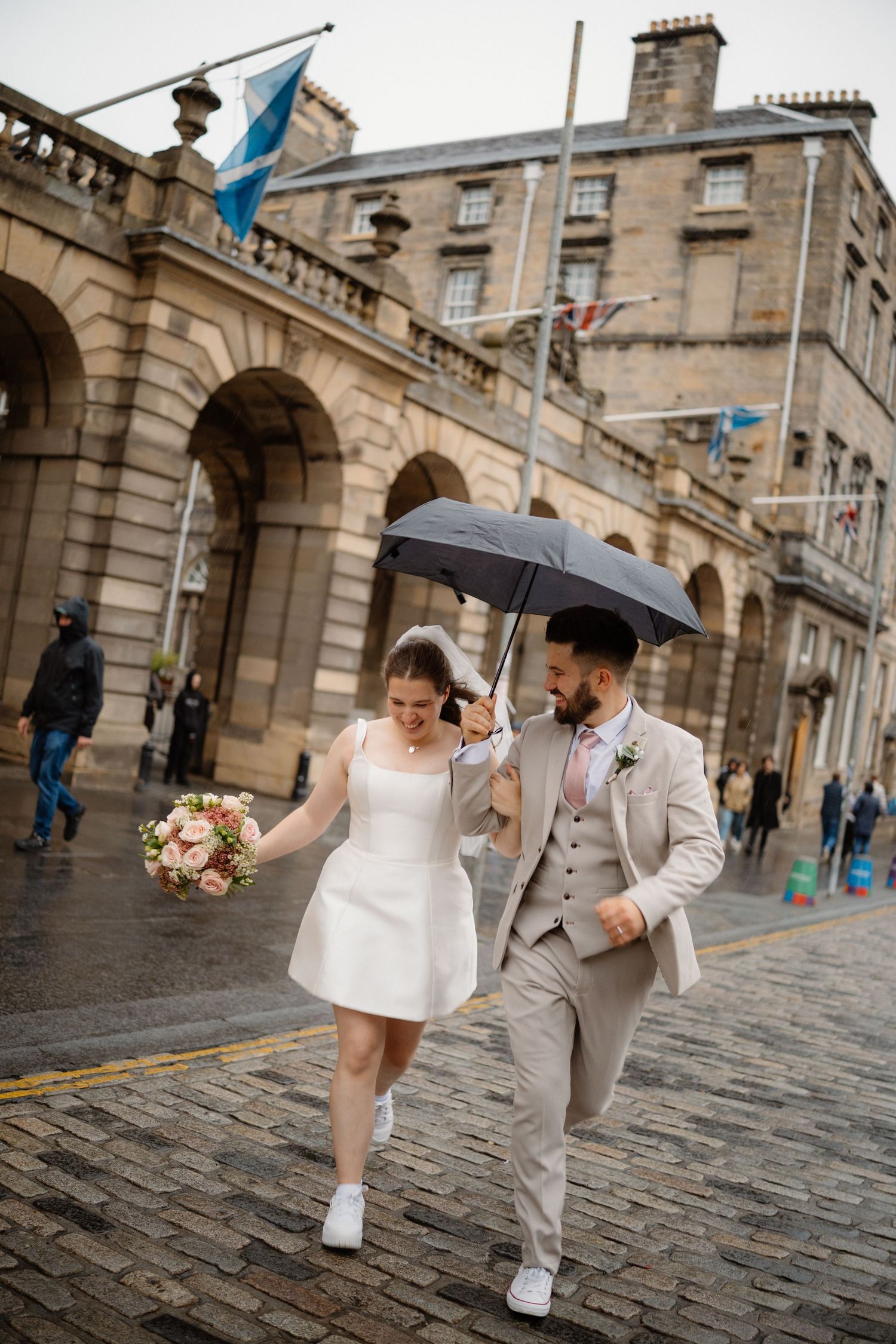 Couple walking together on historic cobblestone street with umbrella and wedding attire in Edinburgh.