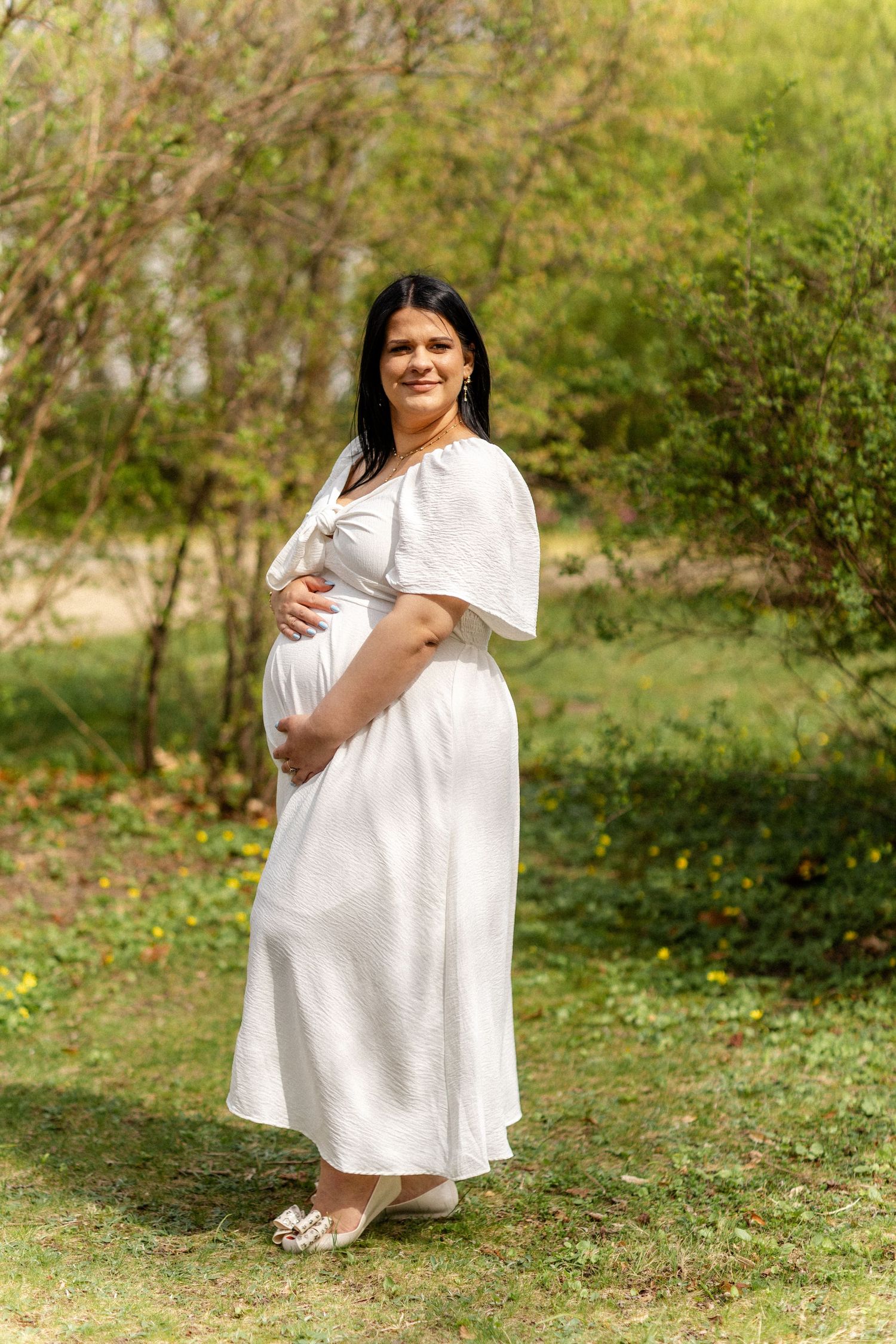 A woman in a white dress poses outdoors in a natural spring setting.