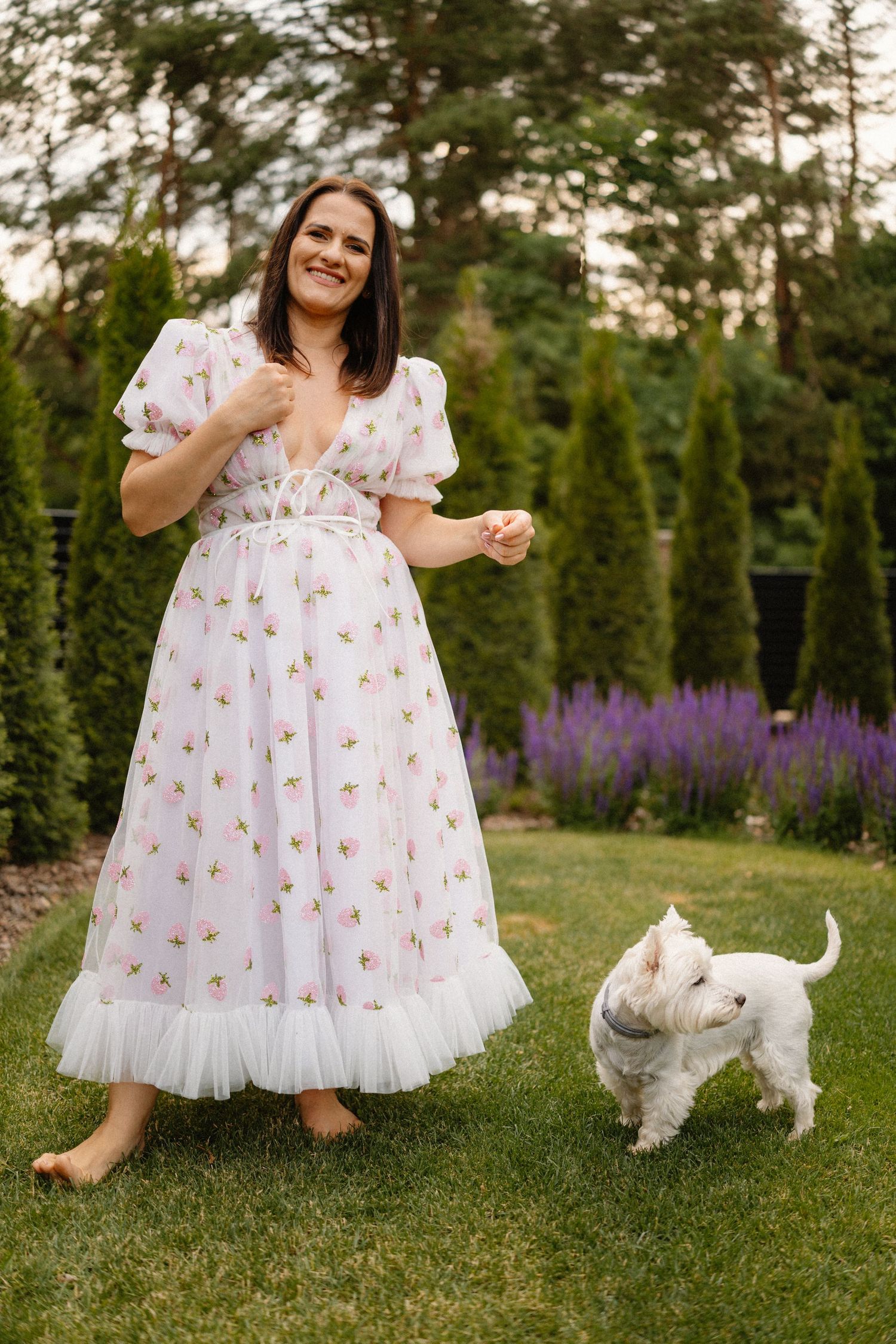 Floral dress details and white dog playing on grass with purple flowers in background.