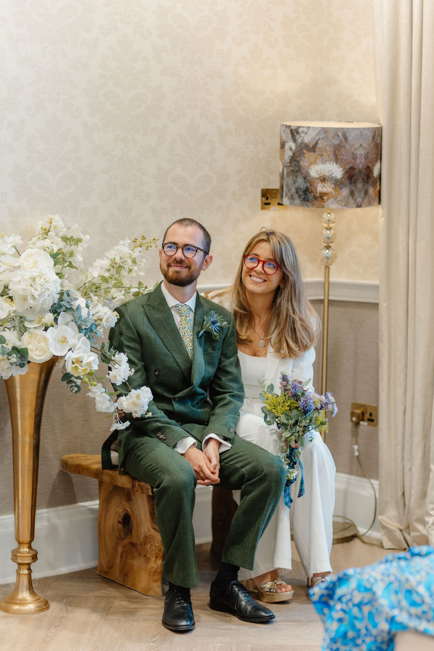 Modern wedding couple poses on wooden bench with gold vases and white floral arrangements.