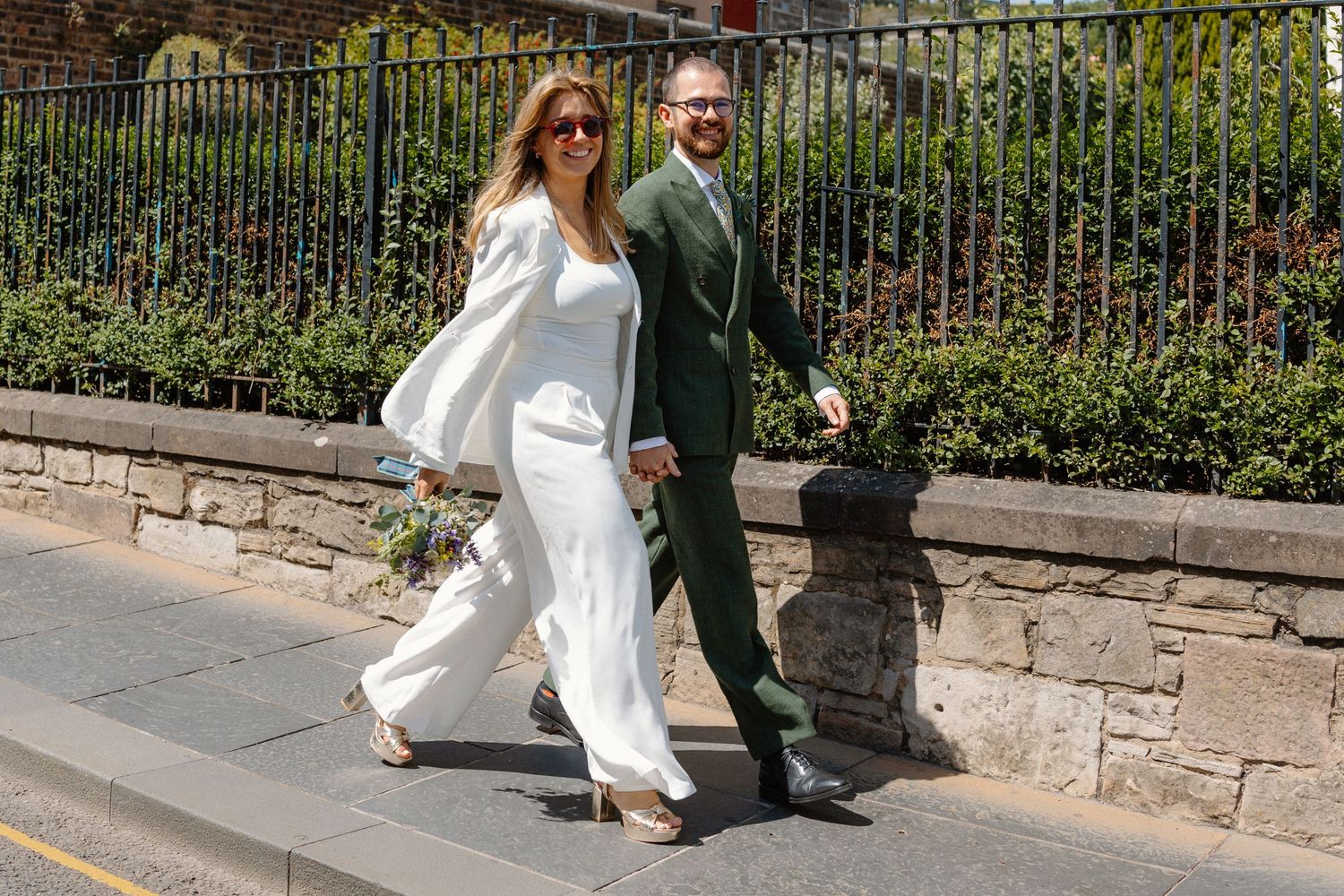 Couple walking along stone wall with ornate iron railing and greenery.