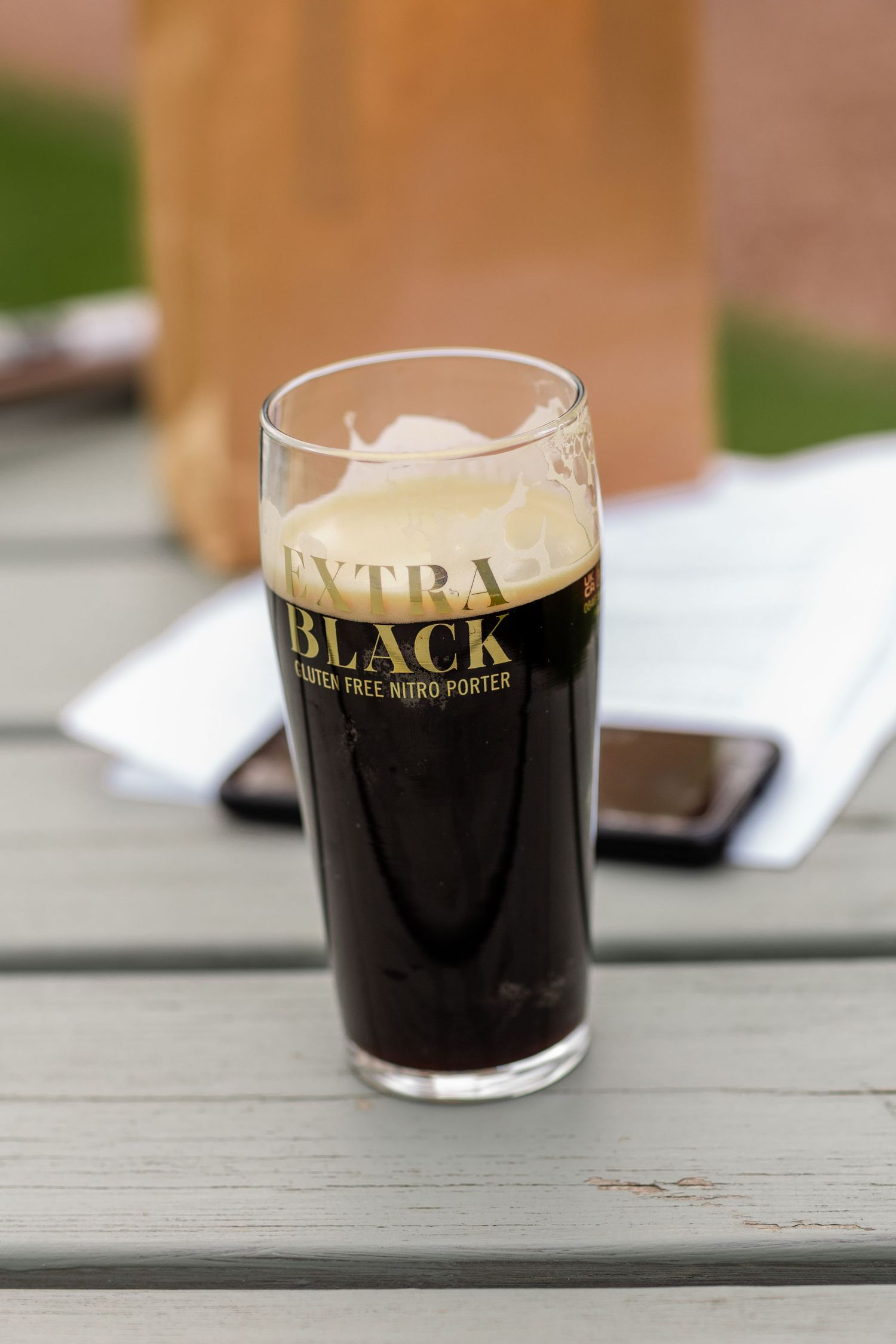 A pint glass of Extra Black gluten-free nitro porter dark beer sits on an outdoor wooden table.