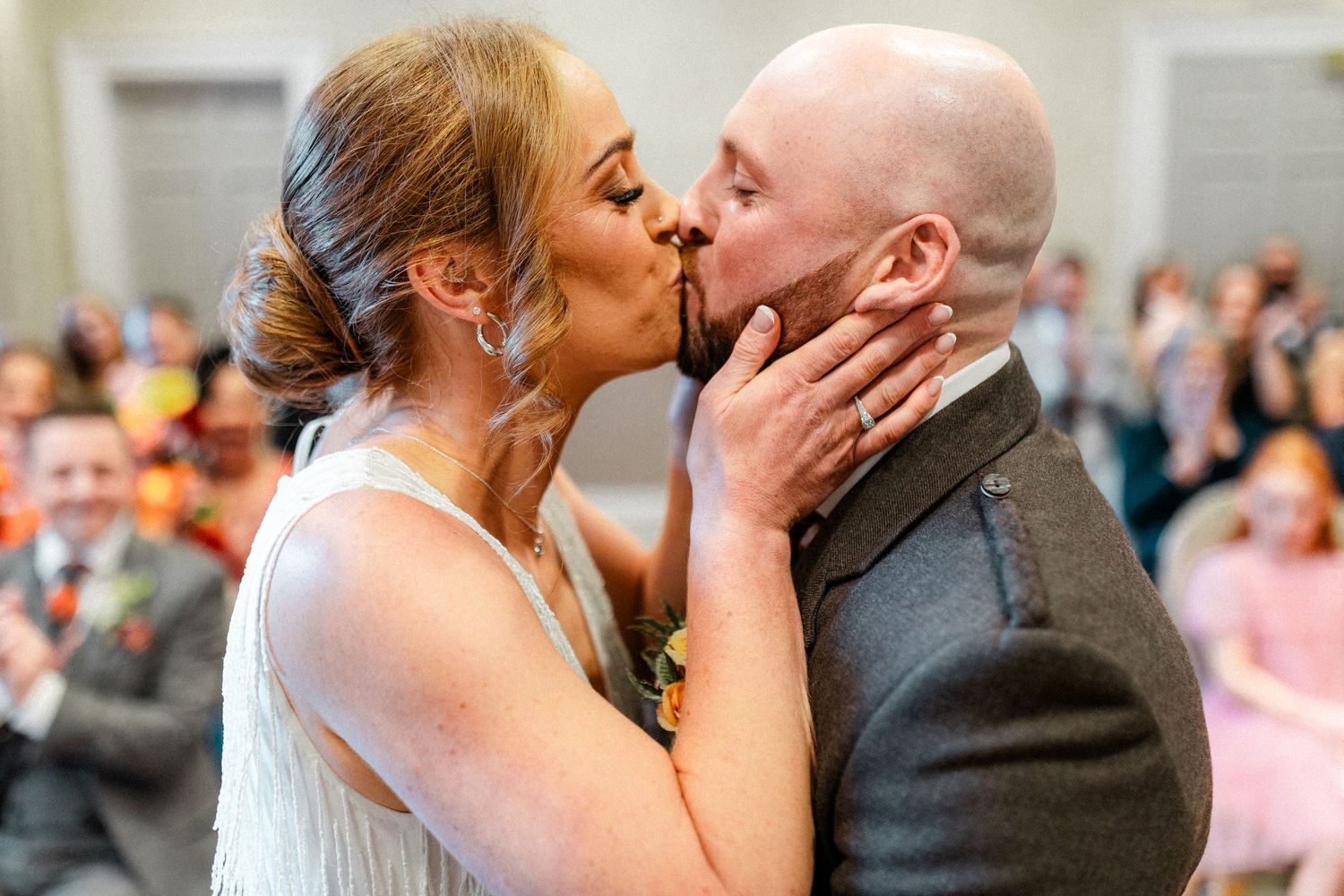 Bride and groom share first kiss at wedding ceremony with guests applauding in background.