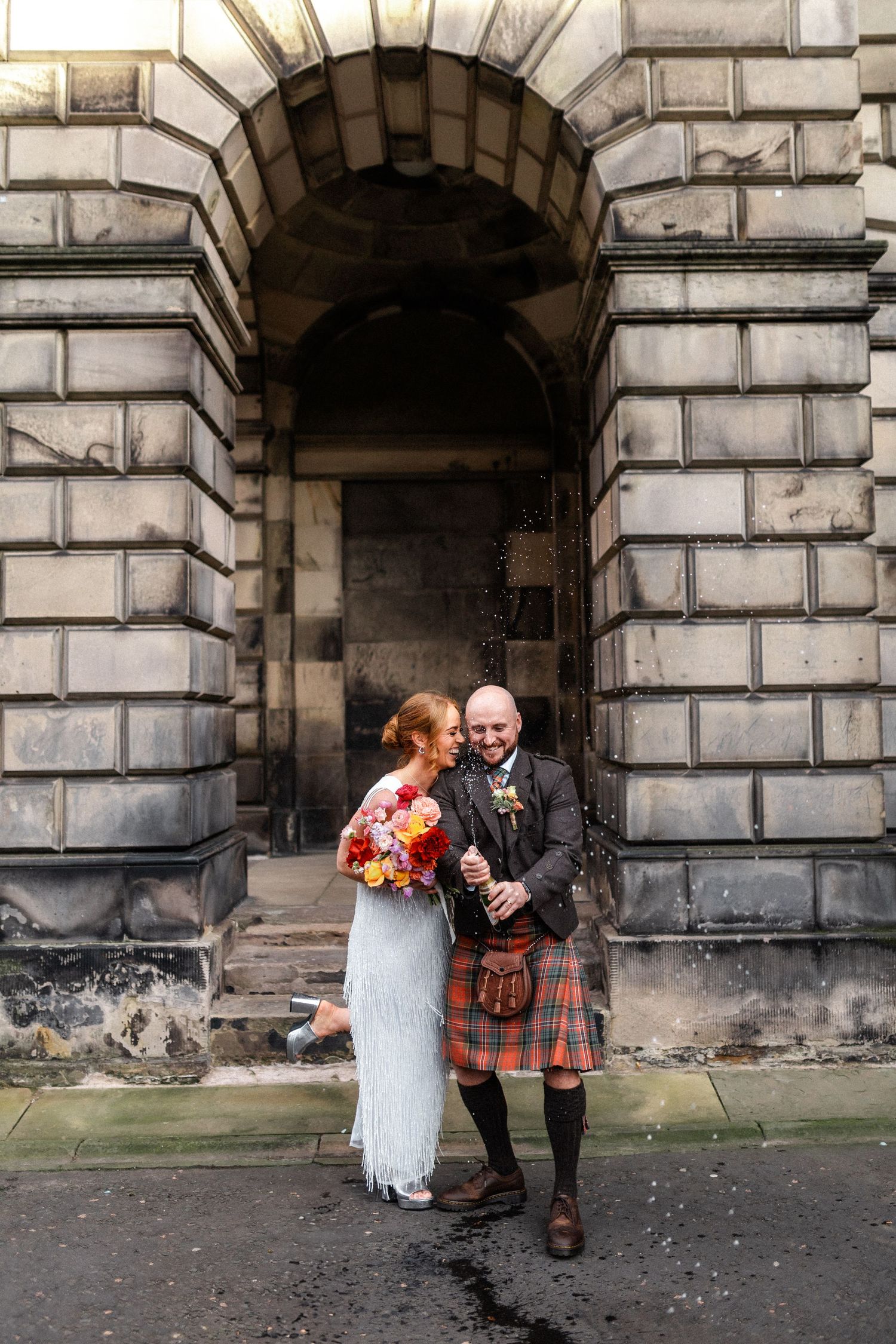 Bride and groom laughing together outside a stone archway, groom wearing a traditional tartan kilt.