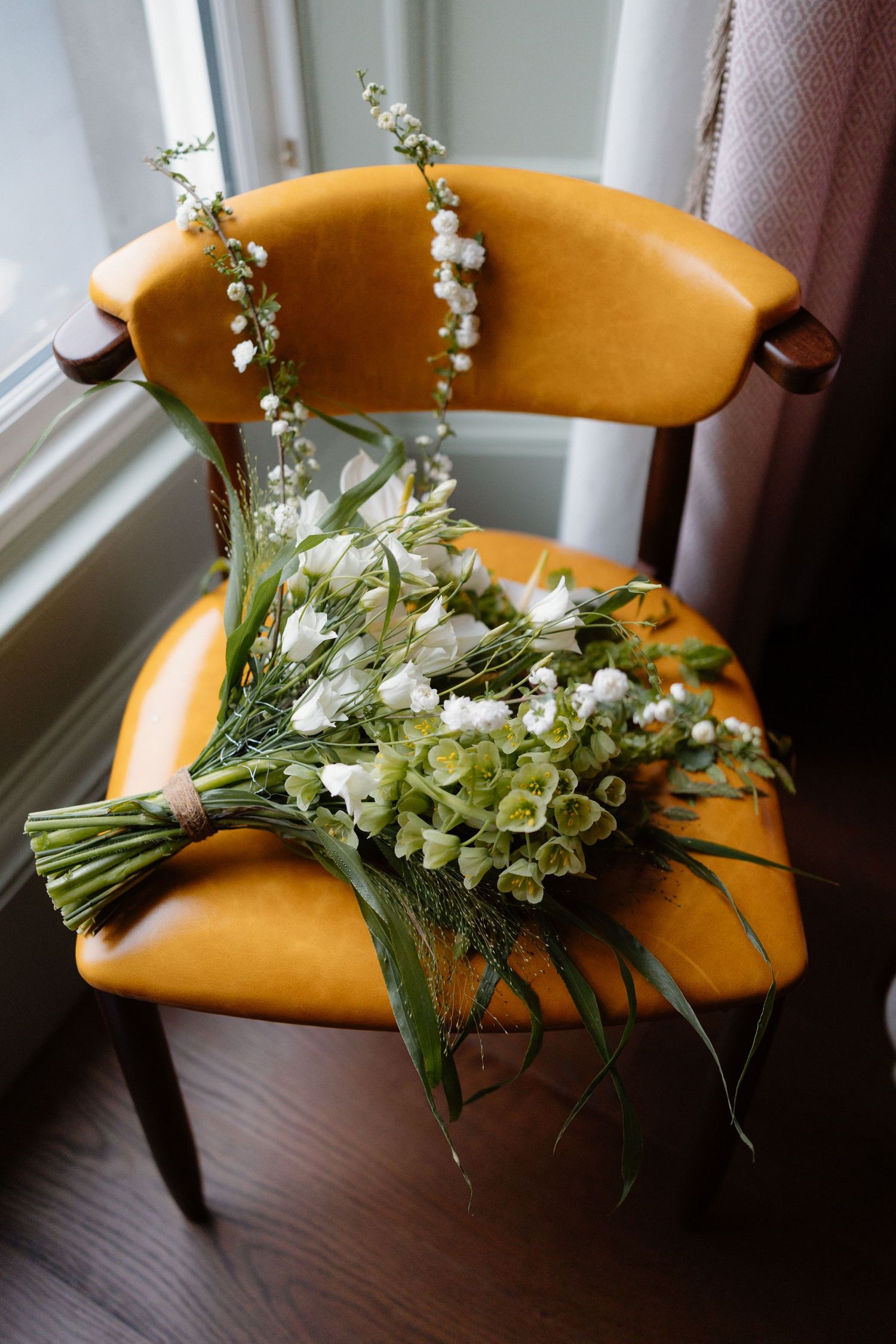 Elegant white and green bridal bouquet with roses and wildflowers resting on a vintage yellow leather chair.