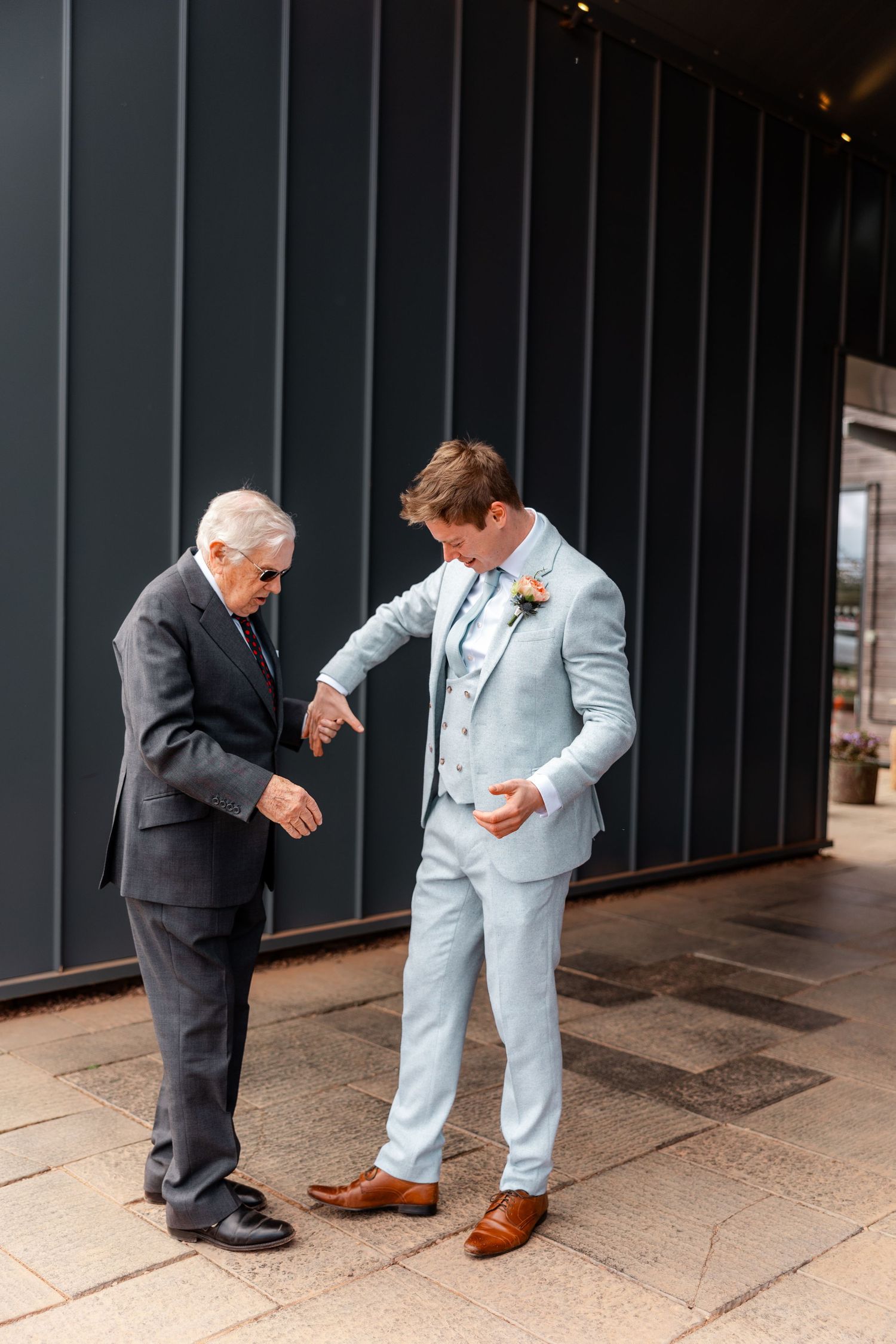 Groom in light blue suit adjusts outfit with older gentleman in dark suit outdoors.