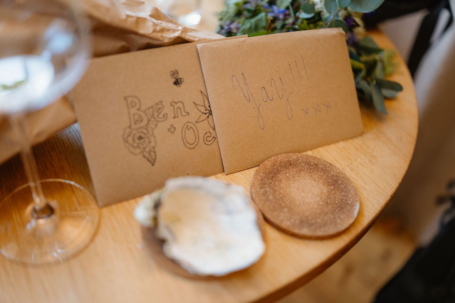 Rustic wedding place cards with floral designs beside an oyster shell and cork coaster on a wooden table.
