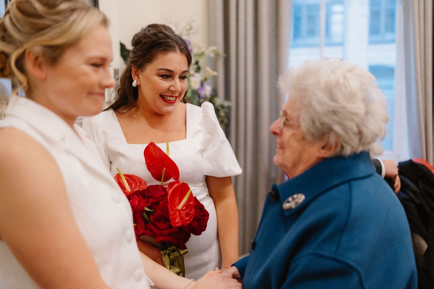 Two brides in white dresses holding red flowers greet an elderly woman in blue at a wedding reception.