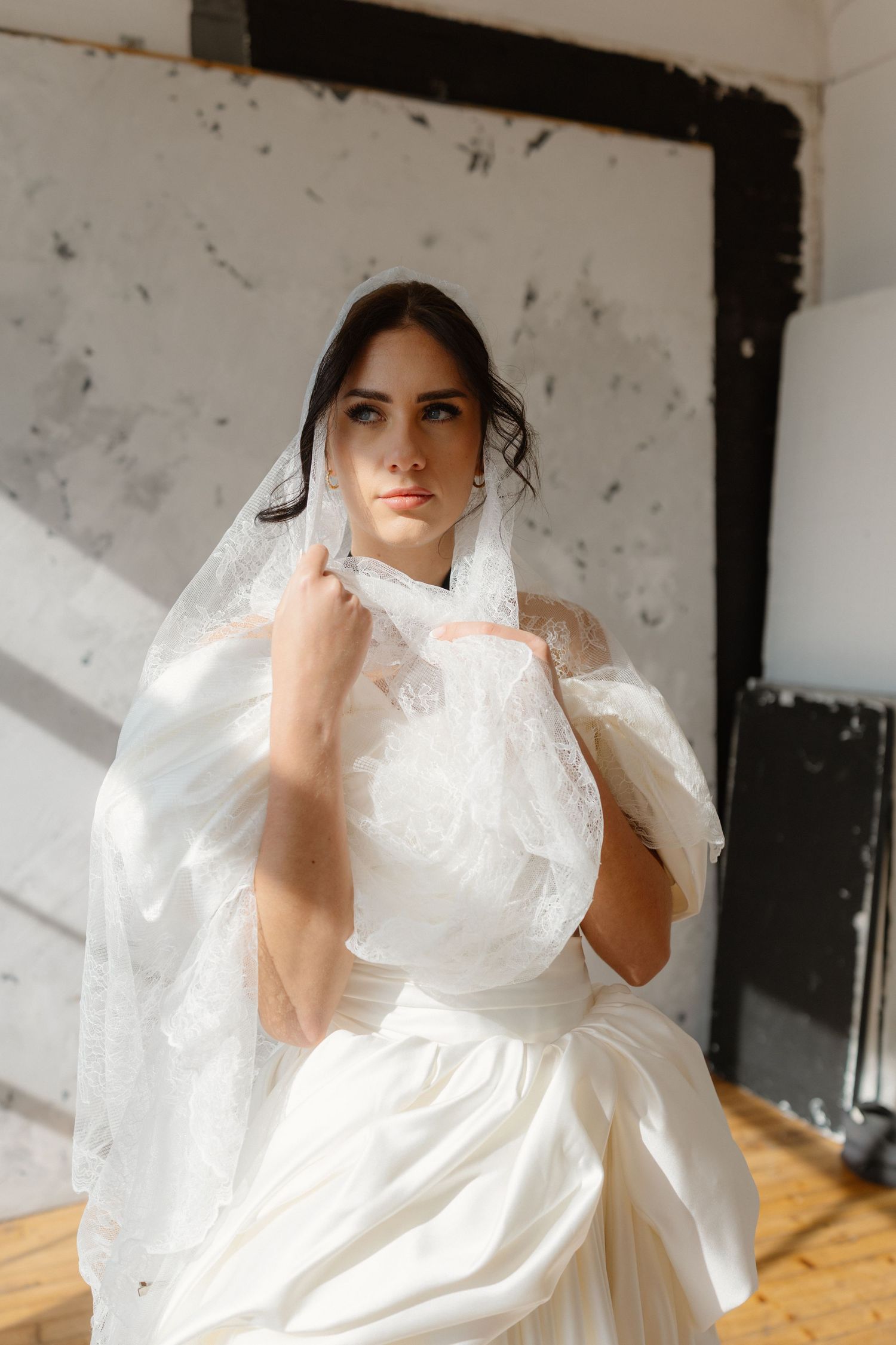 Bride in white satin gown holds lace veil near face, posing in sunlit room with rustic white walls.