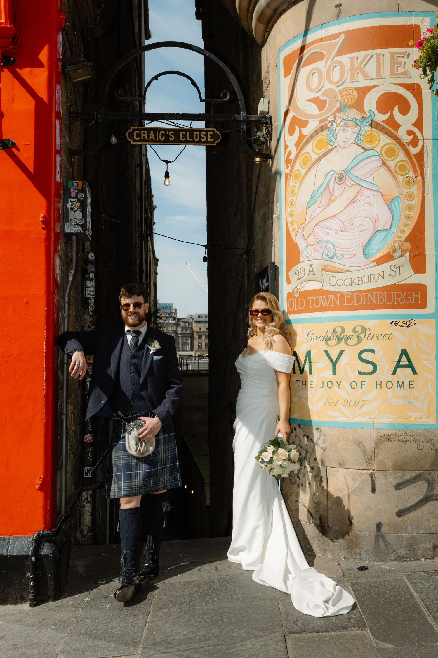 Bride and groom pose at Craig's Close Edinburgh, groom in tartan kilt, bride in off-shoulder gown holding bouquet.