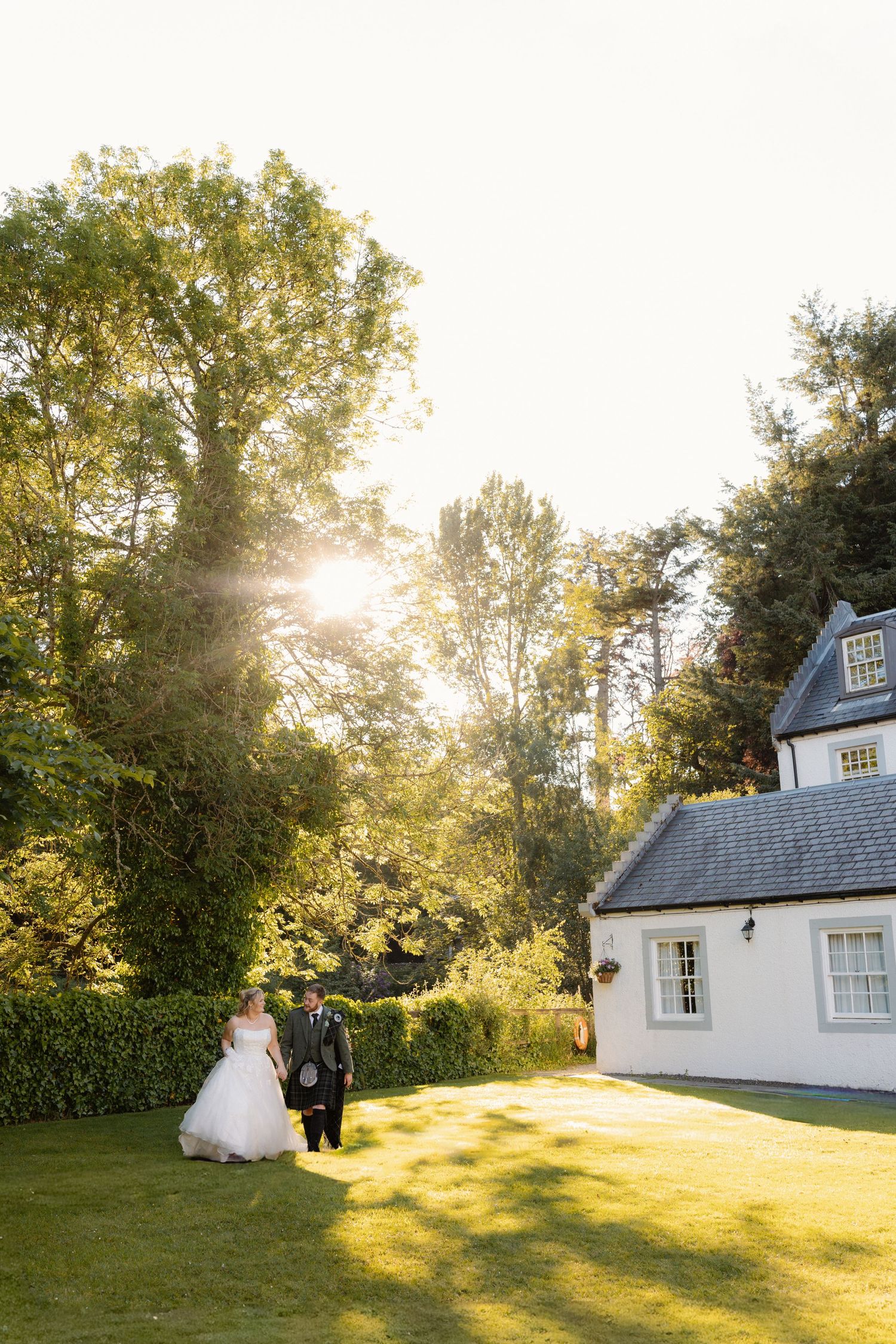 Bride and groom walk hand in hand across sunlit lawn beside a white Scottish country house at golden hour.