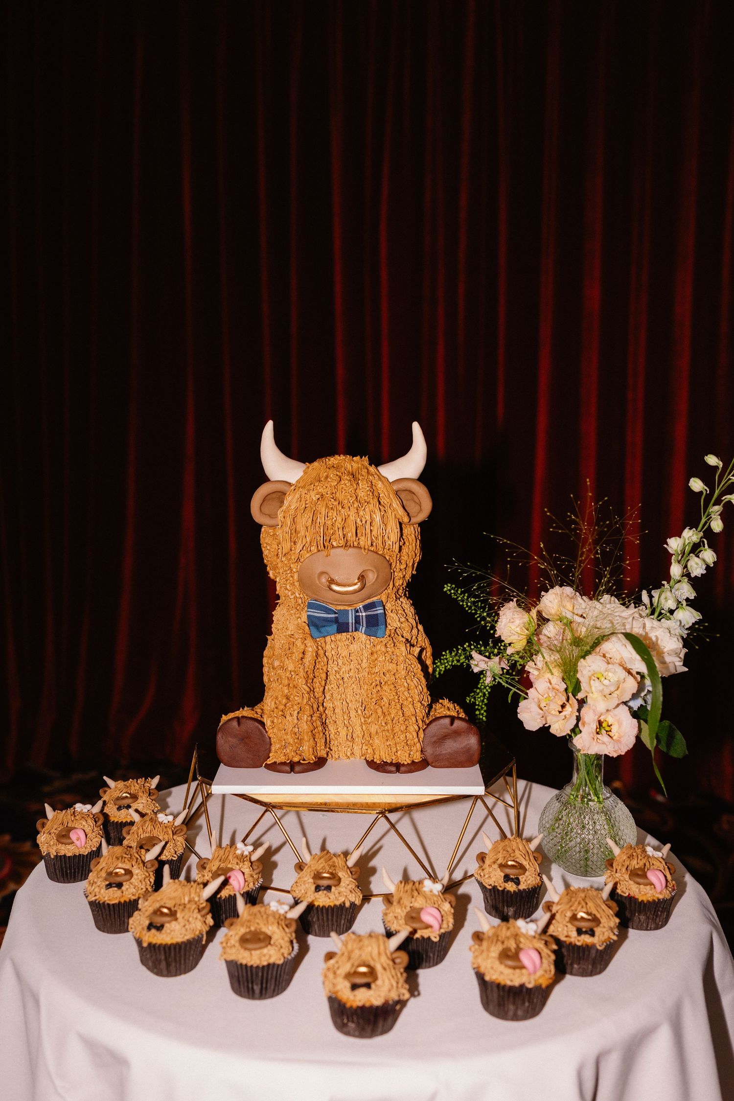Highland cow novelty wedding cake with matching cupcakes on white tablecloth against red curtain.