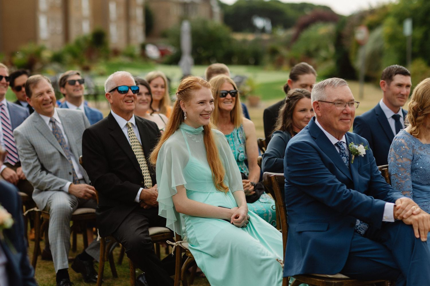 Guests smiling and laughing during an outdoor wedding ceremony, woman in mint green dress seated in the front row.