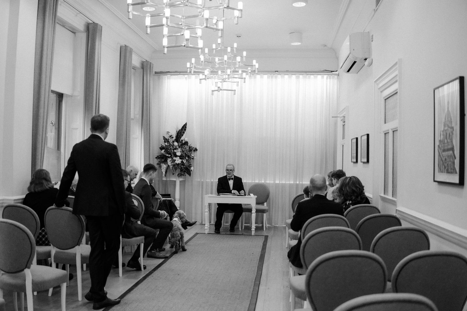 Formal meeting in elegant room with chandelier, suited attendees seated in rows facing a speaker at a desk.