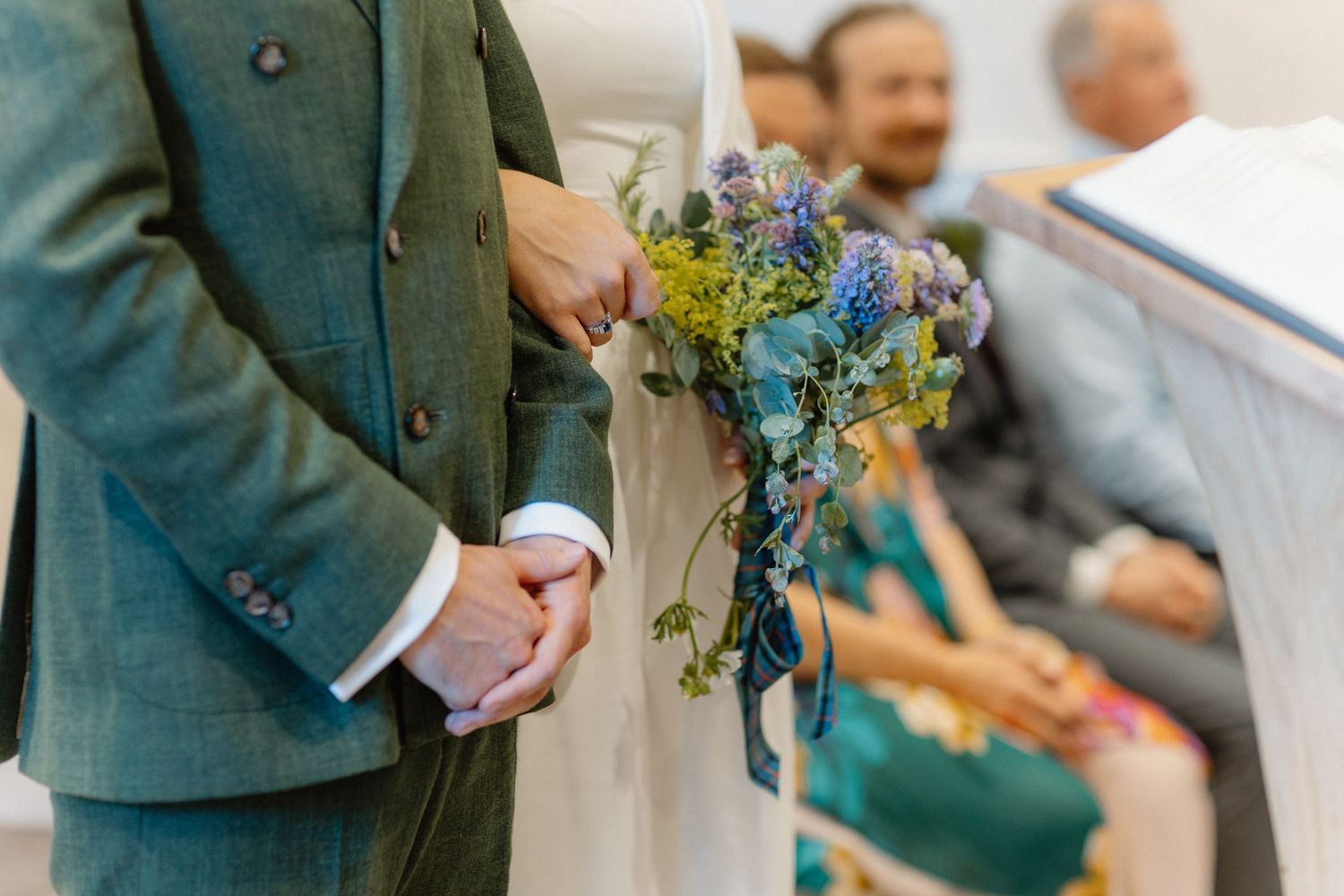 Groom in green tweed suit holds bride's wildflower bouquet during wedding ceremony aisle walk.