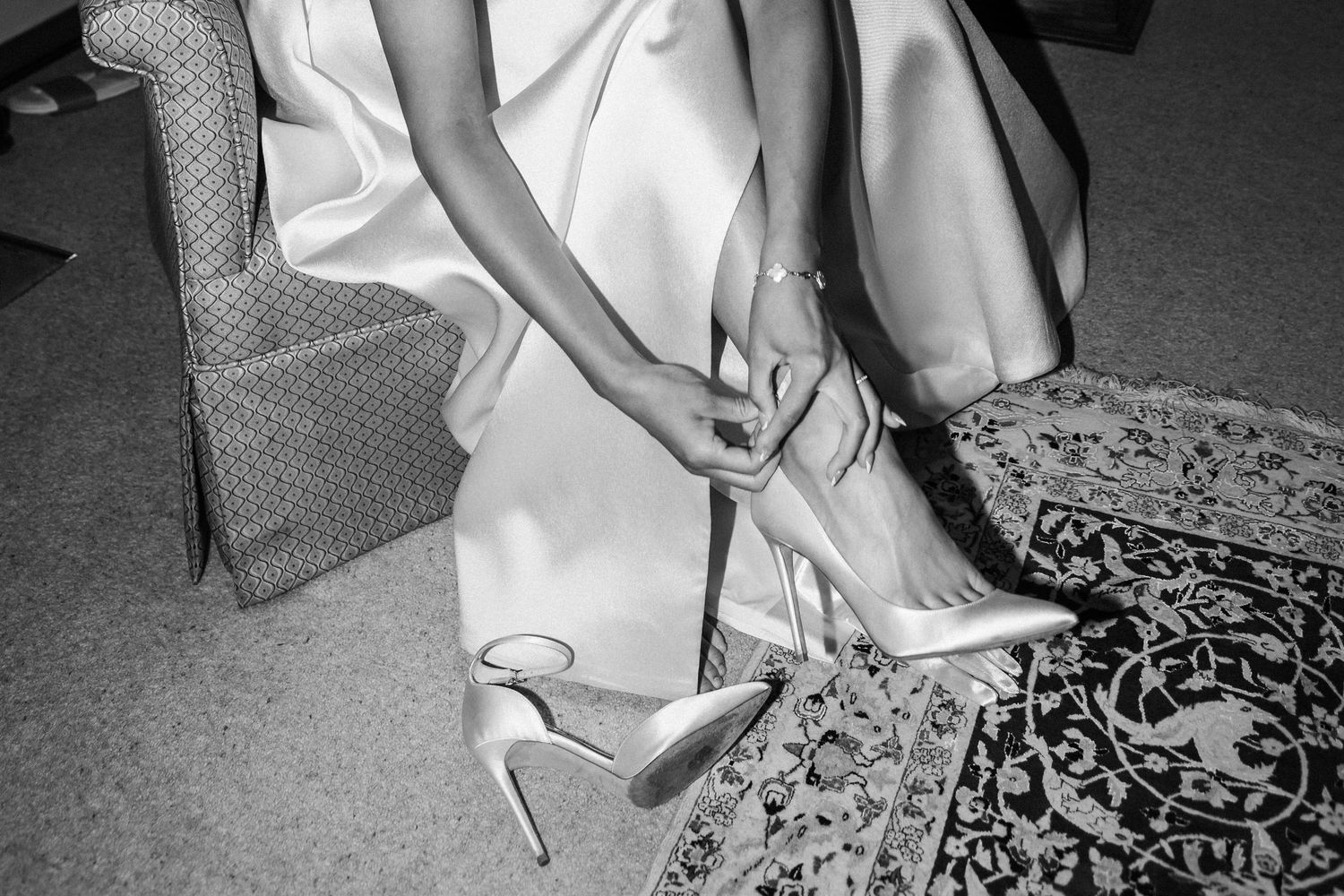 Bride in white gown fastening strappy heels on ornate rug, black and white wedding photography.