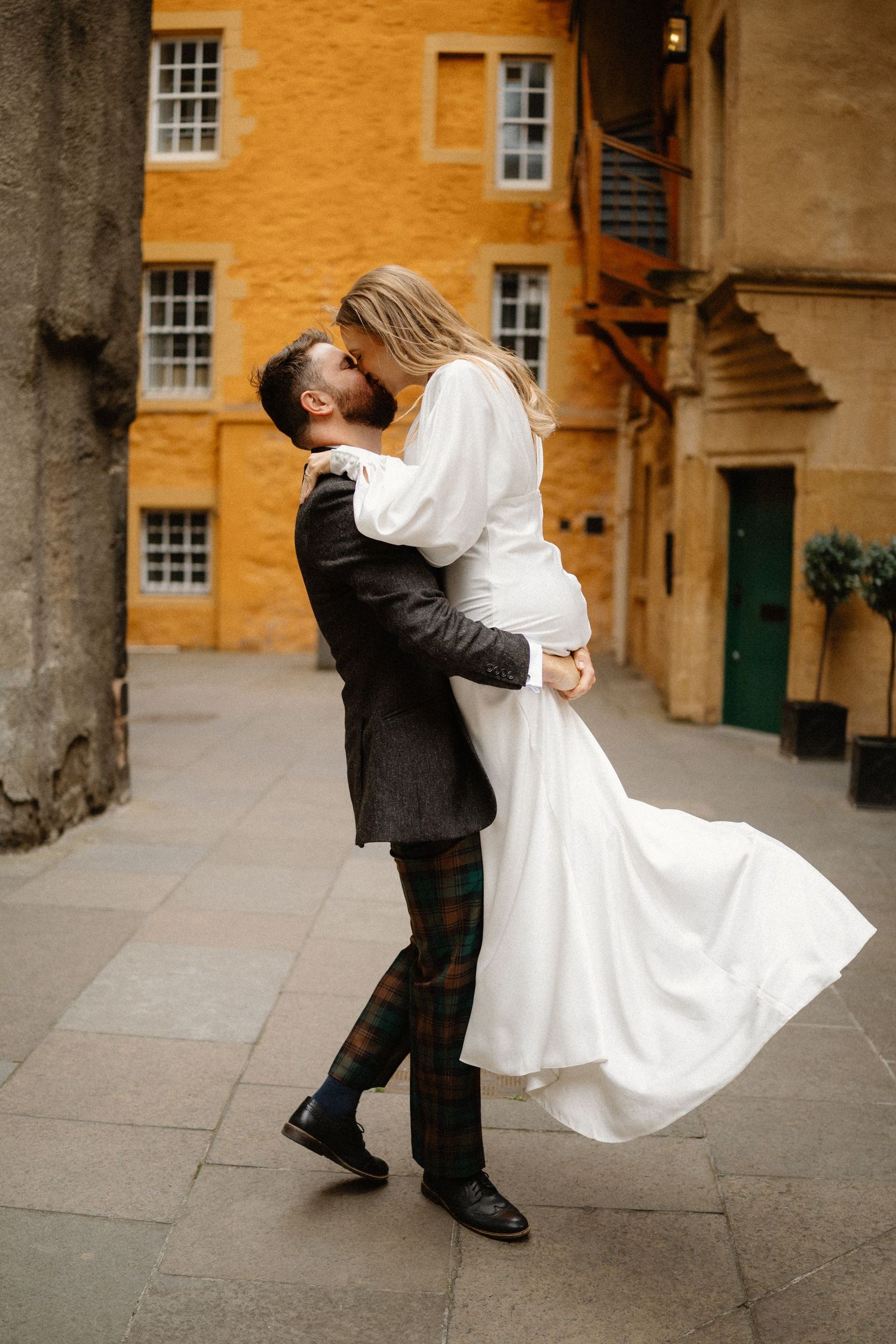 Groom in tartan trousers lifts and kisses bride in white dress outdoors against yellow stone building.