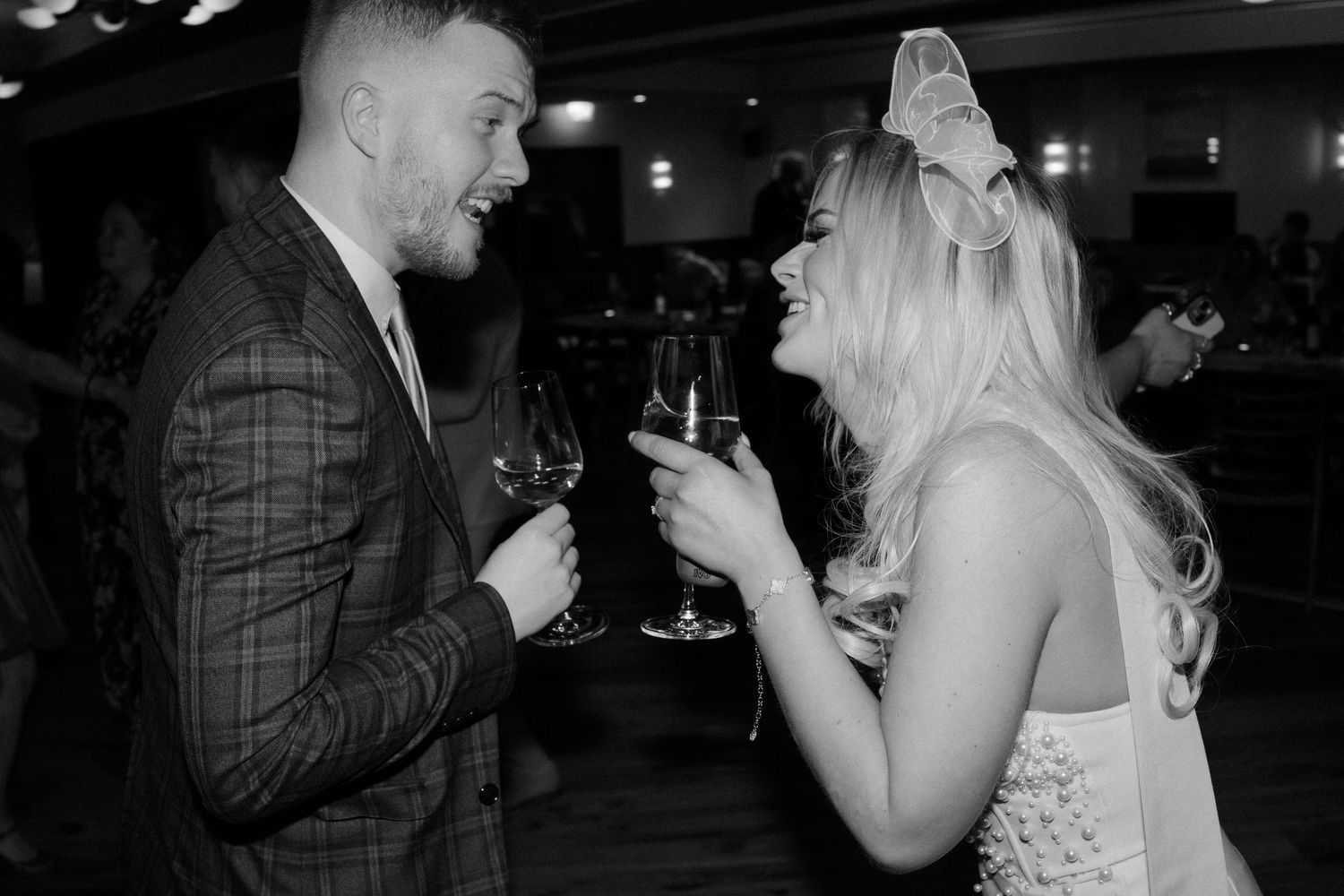 Black and white photo of a laughing bride and groom toasting with wine glasses at their reception.