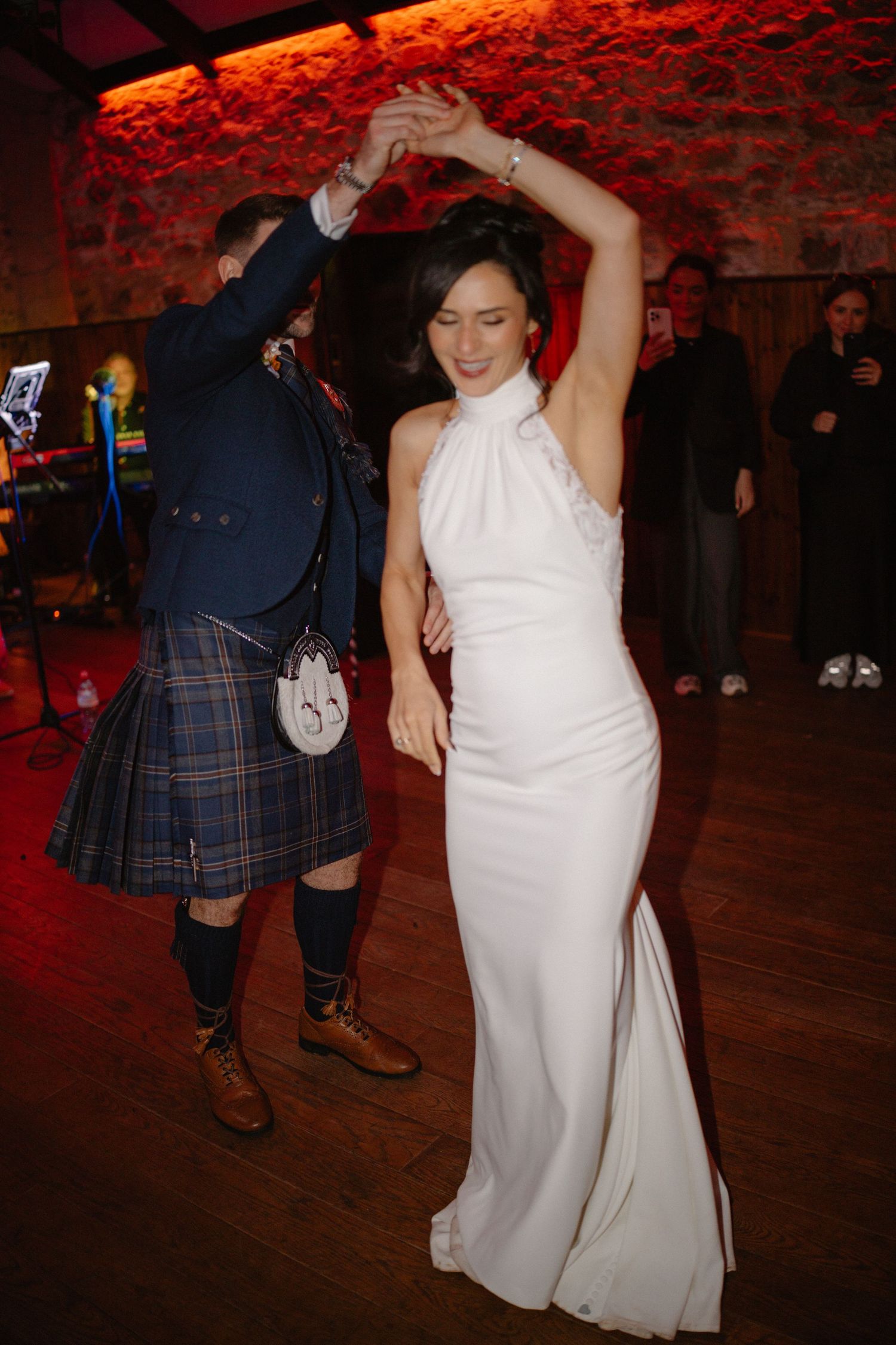 Bride in white halter gown dancing with groom in Scottish kilt at wedding reception.