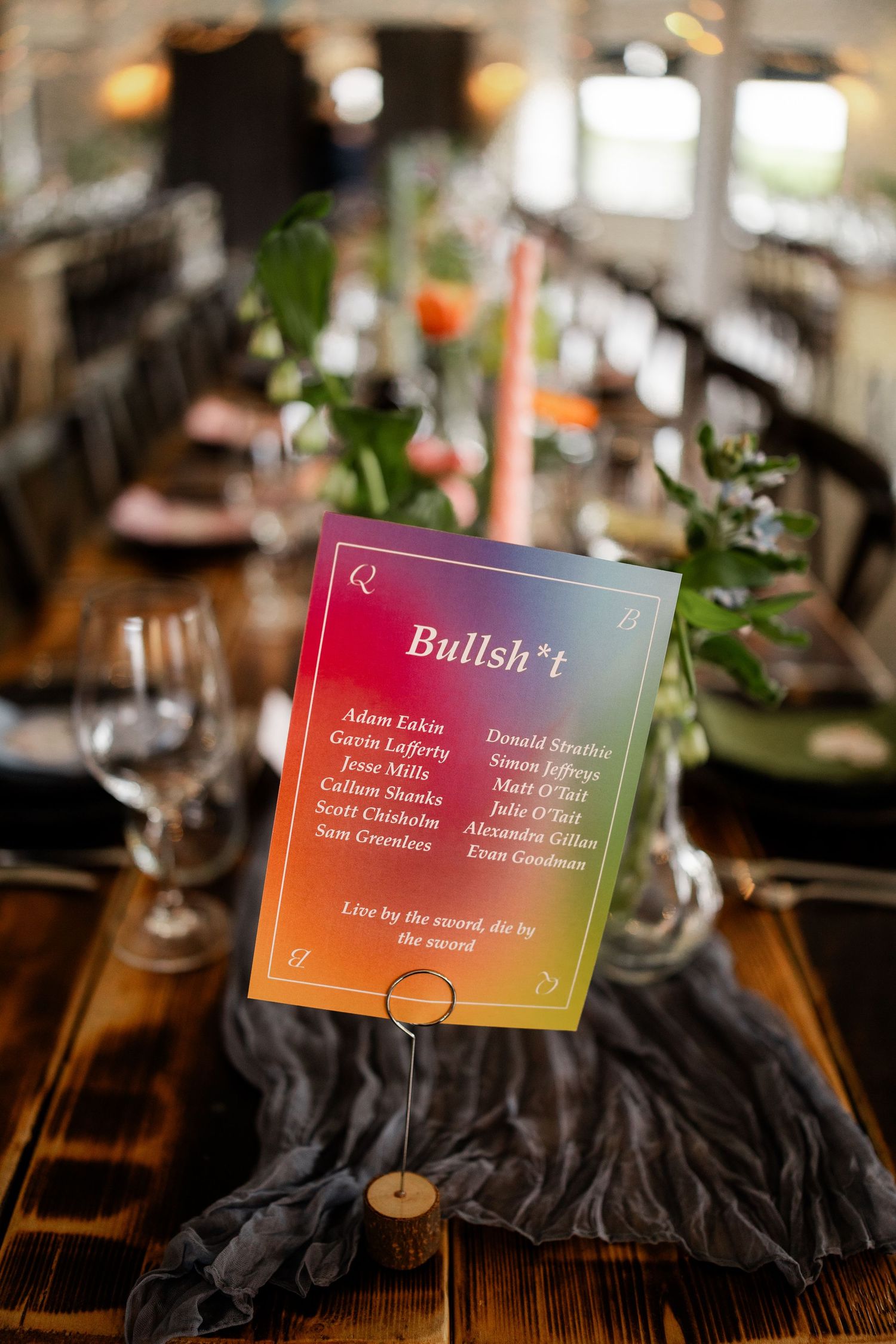Colorful gradient wedding table sign reading Bullsh*t with guest names on a rustic wooden reception table.