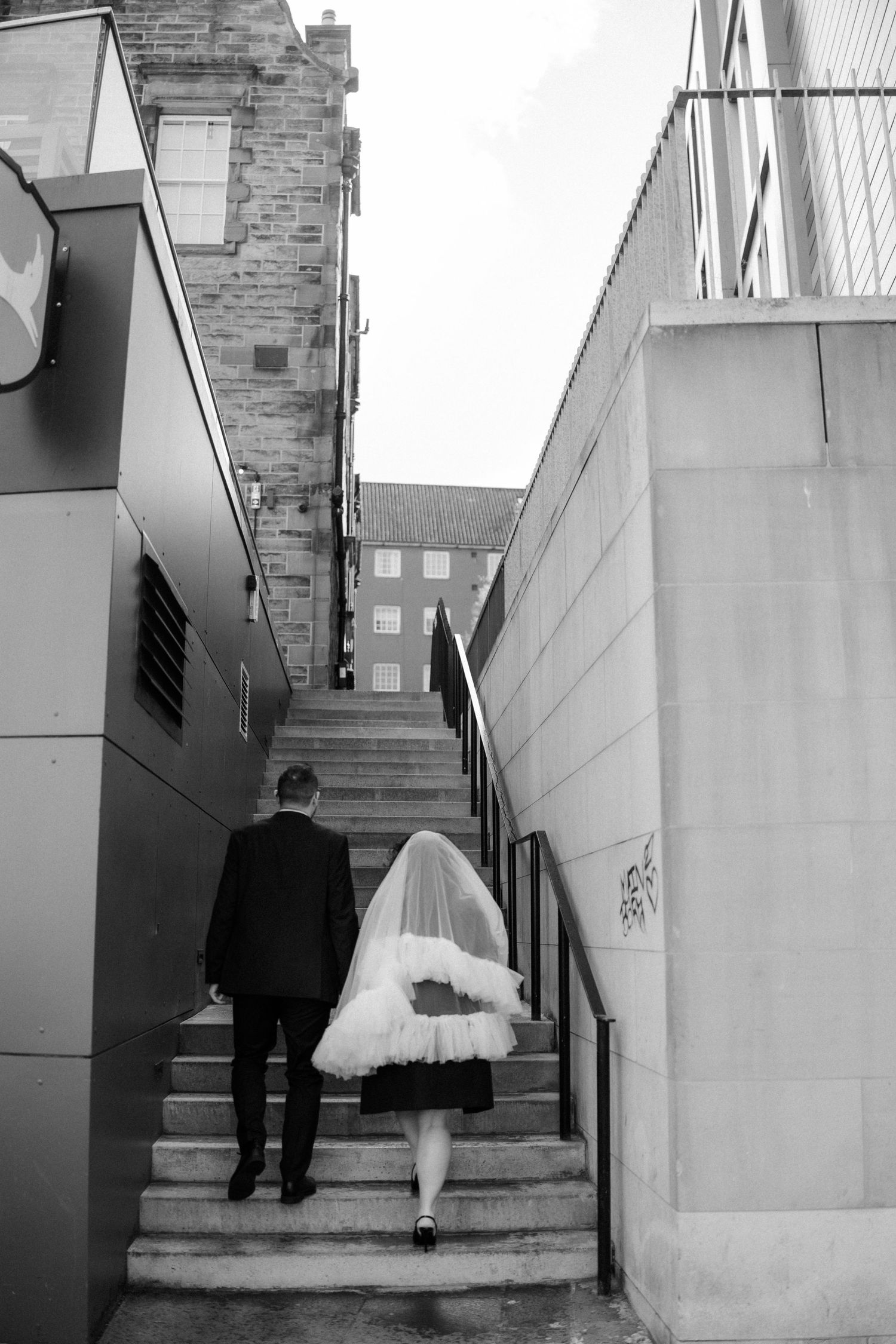 Black and white photo of bride and groom walking up outdoor stone steps between modern buildings.
