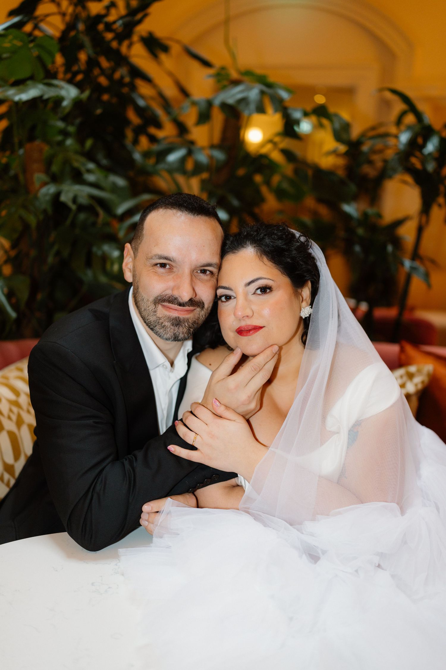 A bride and groom pose together at a table, she wears a veil and red lipstick, he wears a black suit.