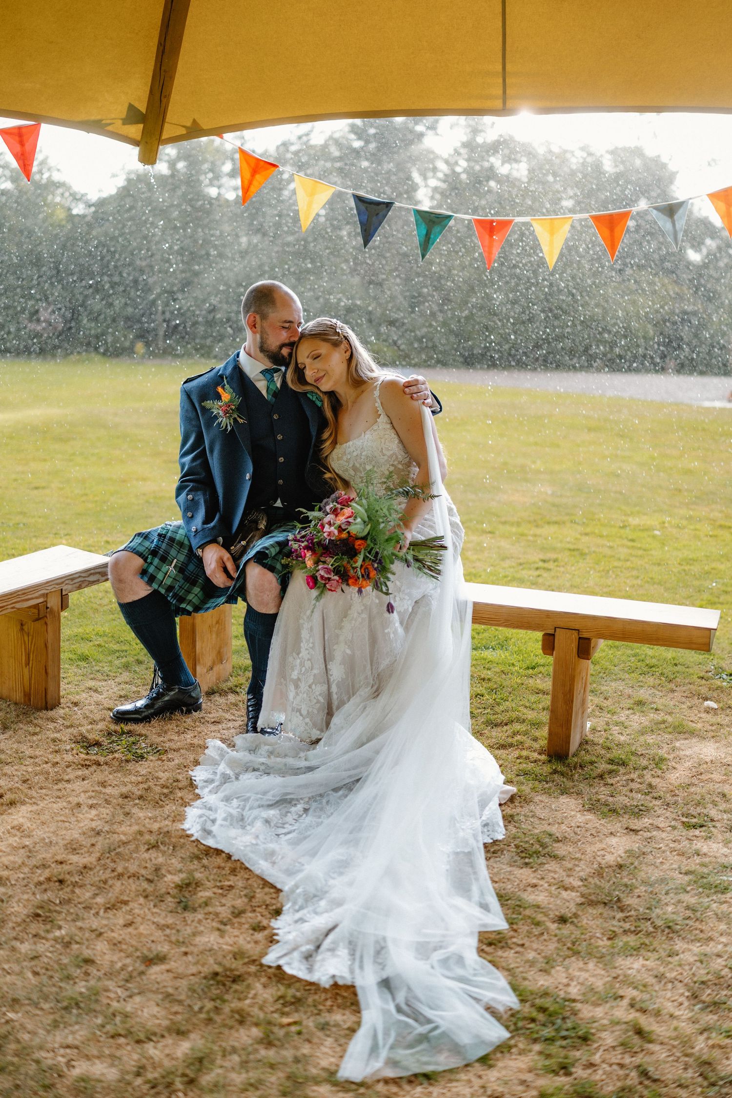 Bride and groom sit together under colorful bunting shelter in the rain, bride's lace gown flowing gracefully.