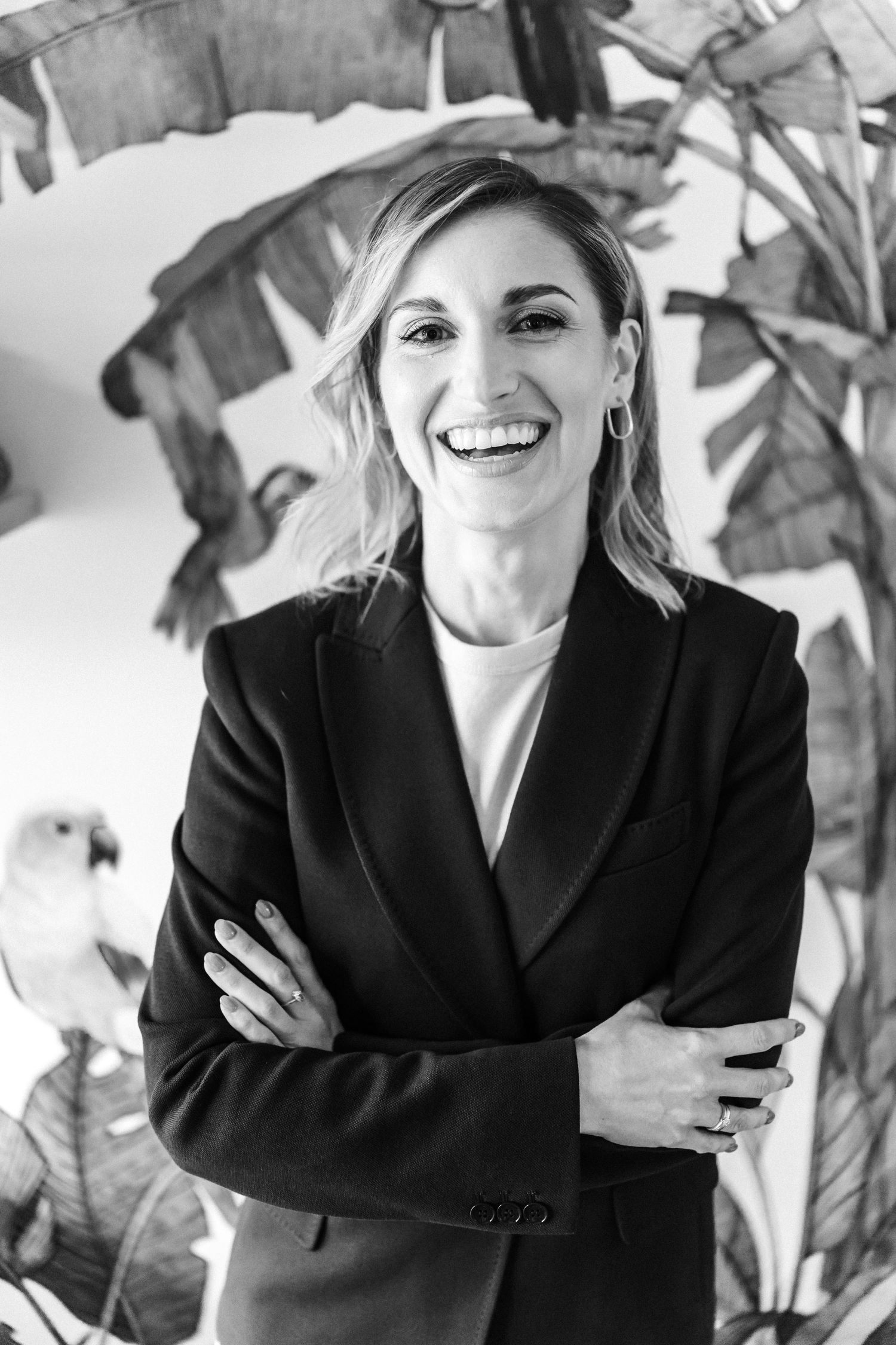 Professional black and white portrait of a person in a black blazer smiling against a tropical leaf background.