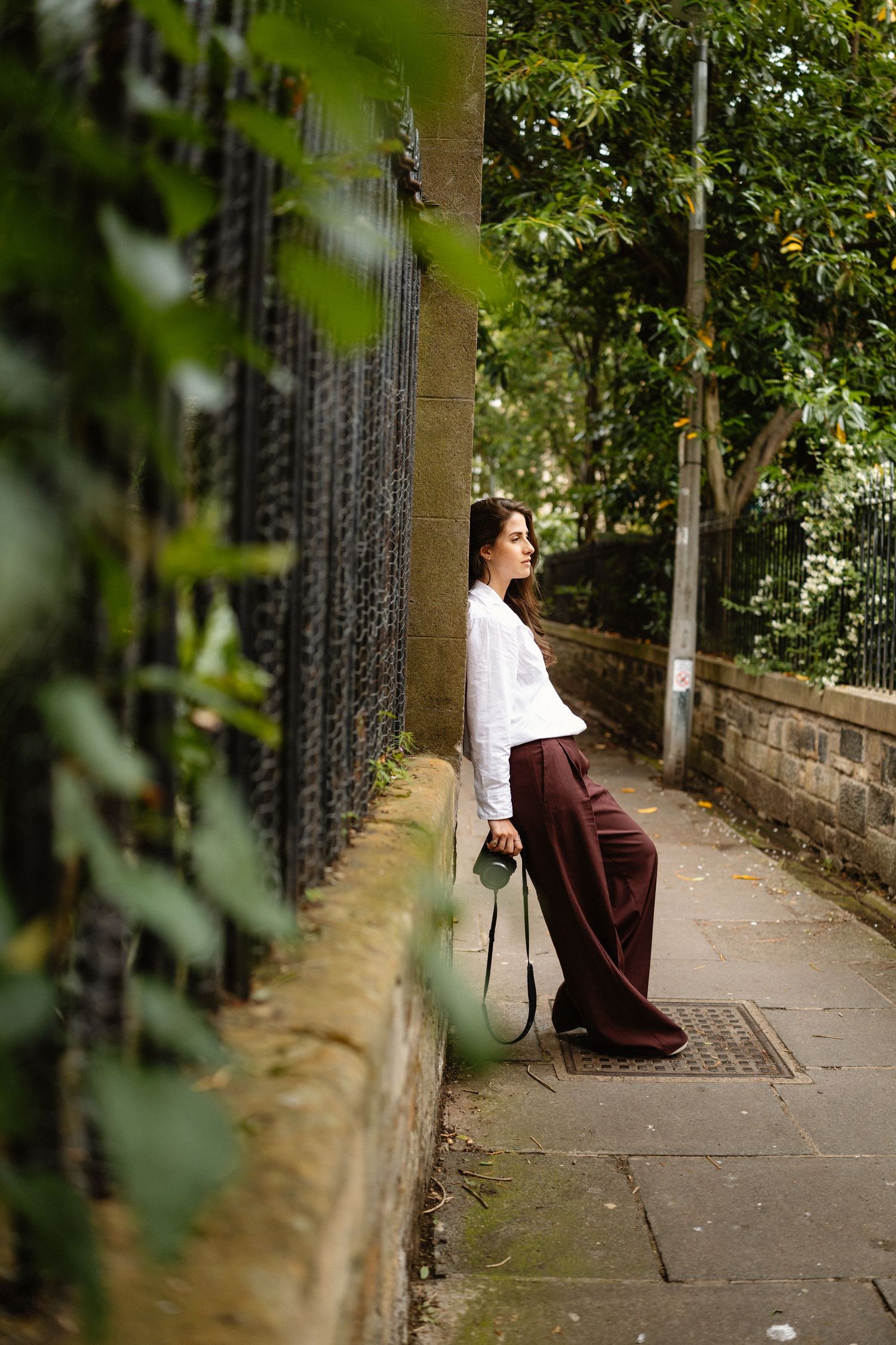 Model in white and burgundy outfit sits on stone wall near greenery.