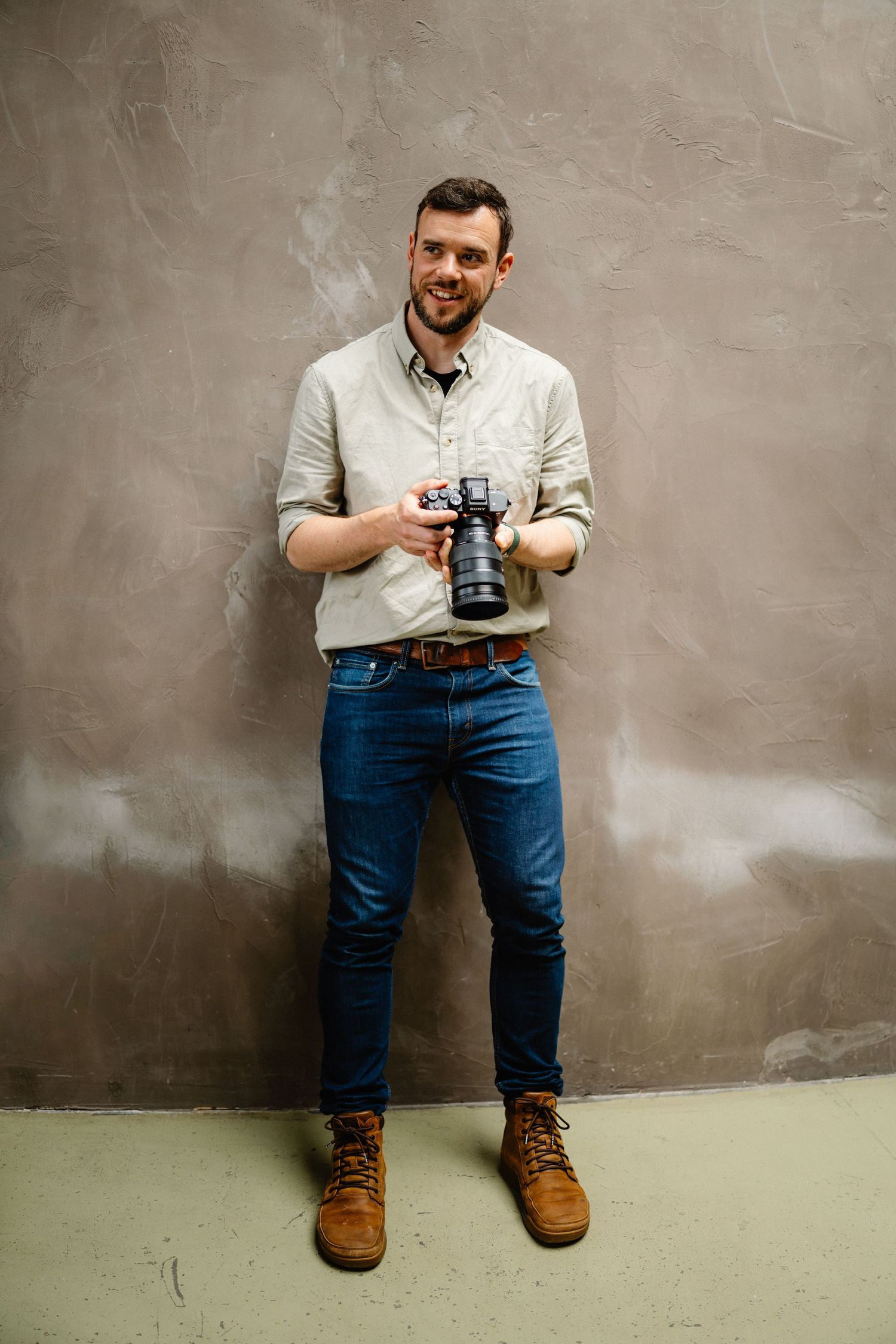 Photographer in casual attire holds professional camera against grey backdrop.