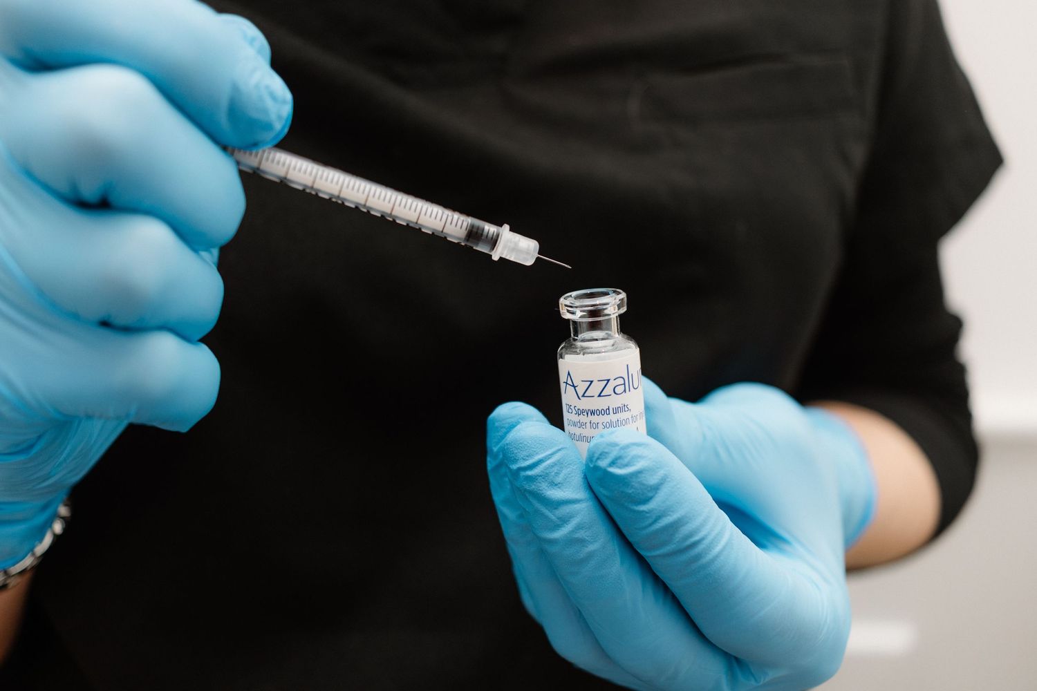 Hands wearing blue medical gloves holding syringe and vial in clinical setting.