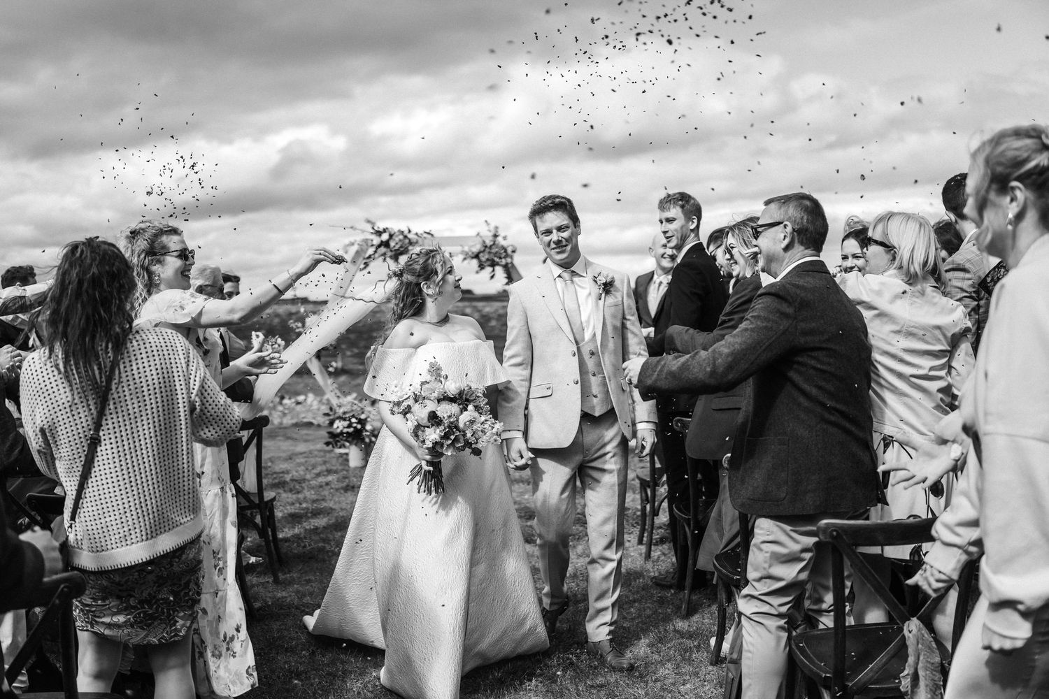 Black and white photo of wedding guests throwing confetti during outdoor celebration.