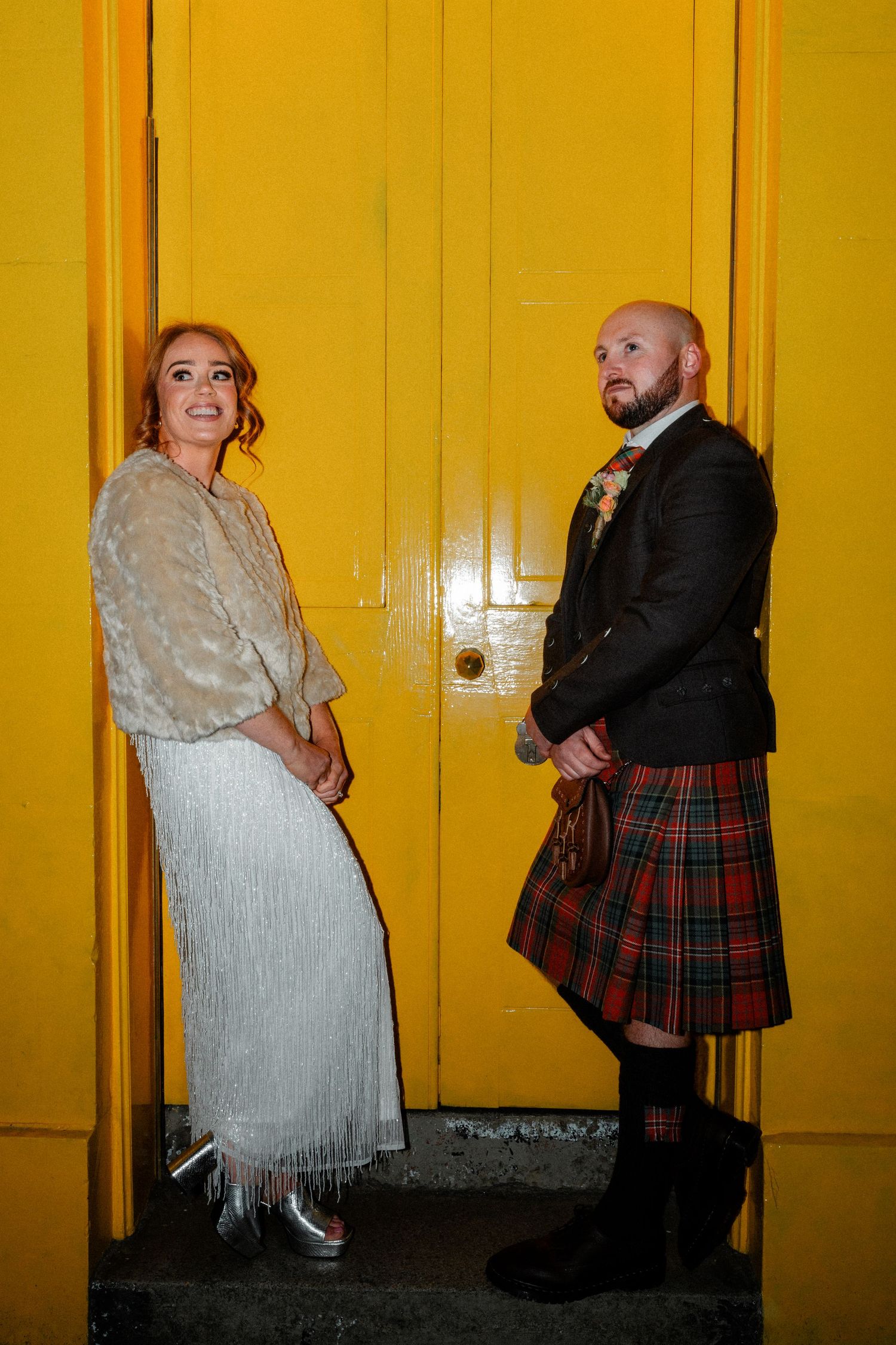 A couple poses against a bright yellow wall, with one wearing a traditional Scottish kilt.