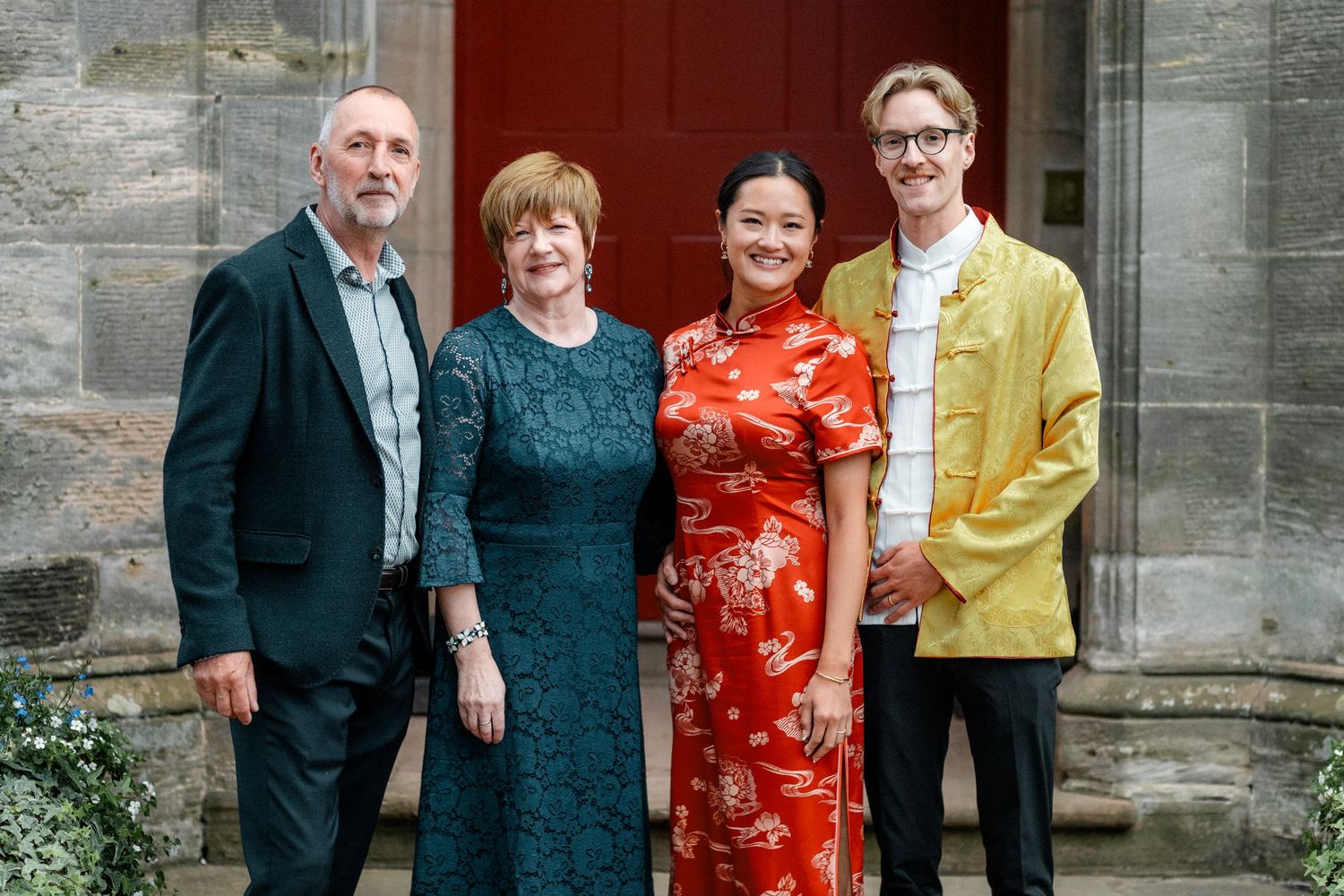 Four people in formal attire pose together against a stone building backdrop.