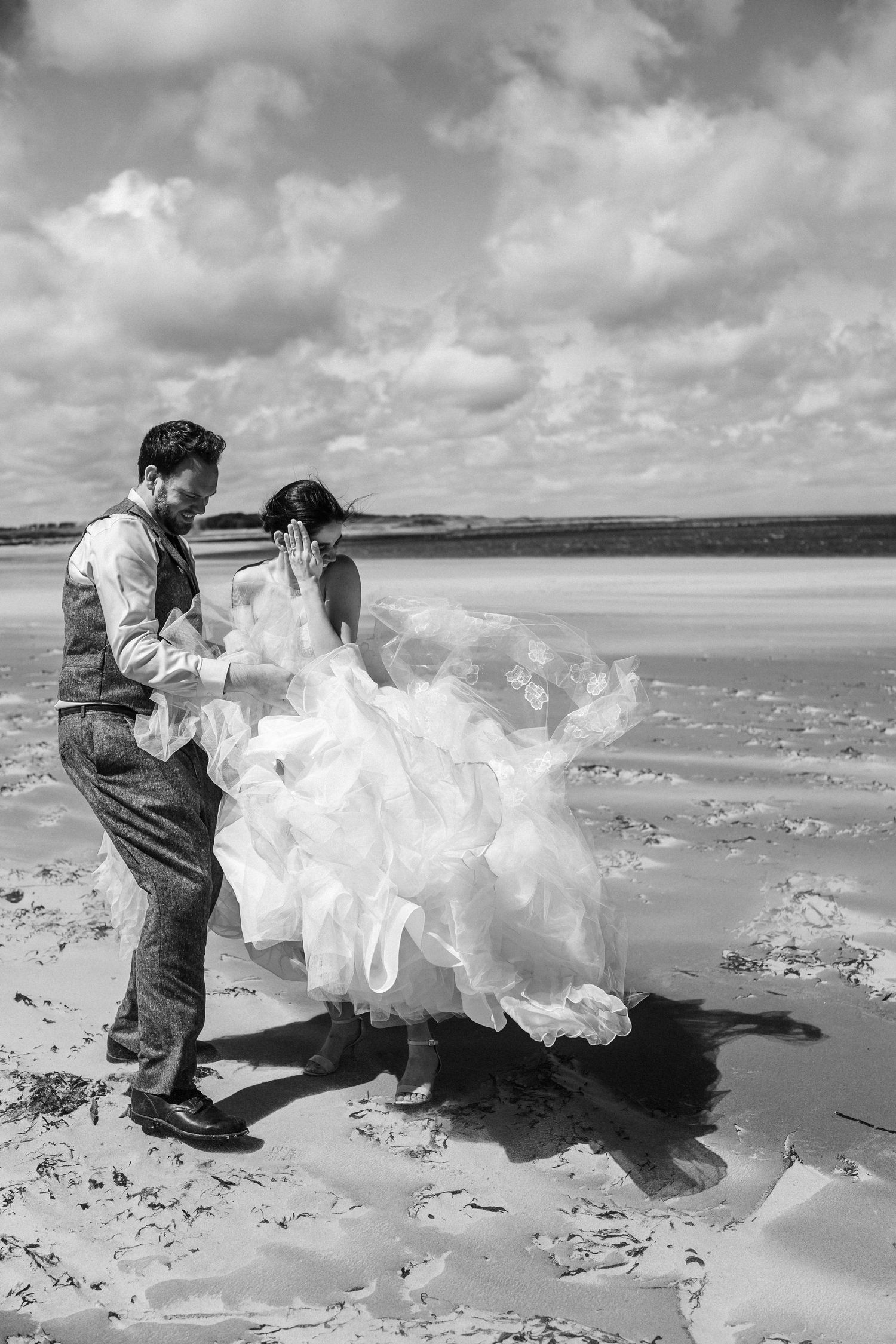 Dynamic black and white photos of a couple playfully posing on a windswept beach.