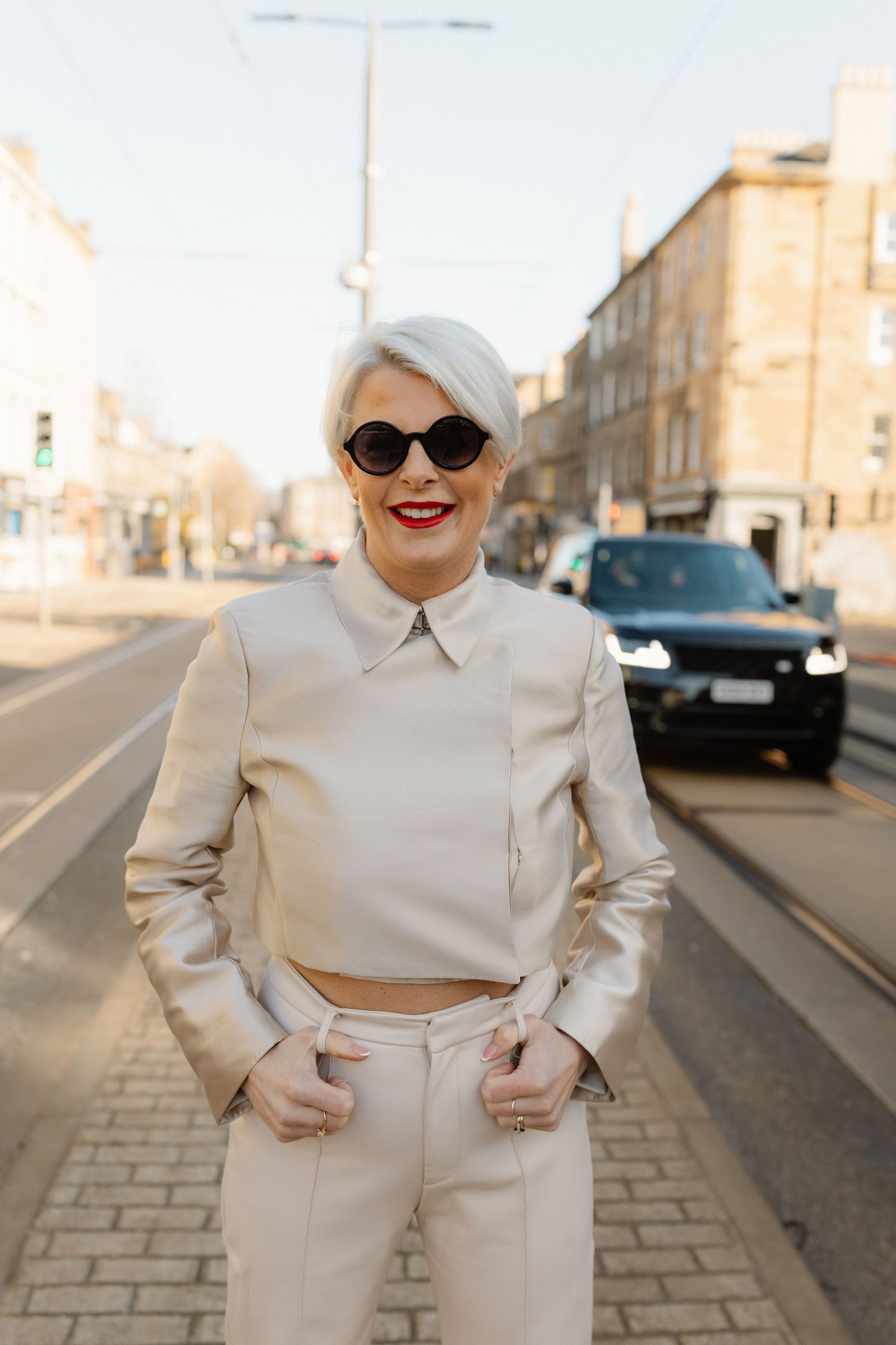 A person poses in an all-white outfit with sunglasses and red lipstick on a city street at sunset.