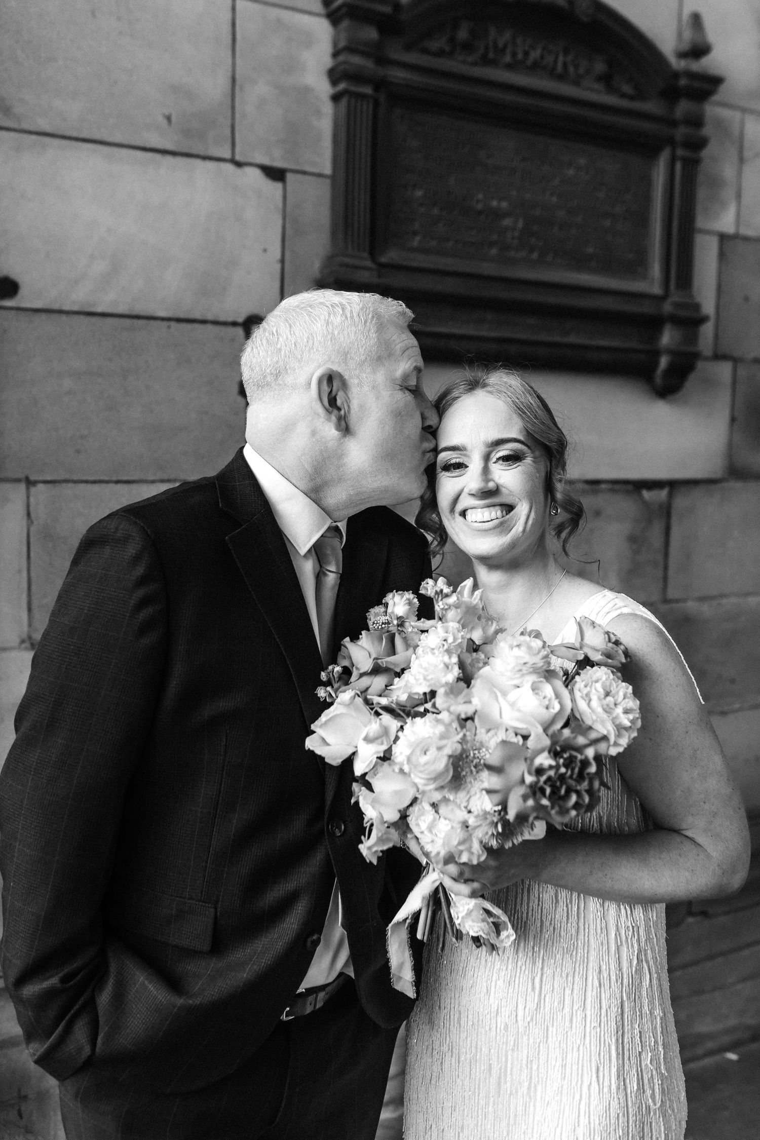Black and white photo of a smiling bride holding bouquet receiving a kiss on cheek from older man in suit.