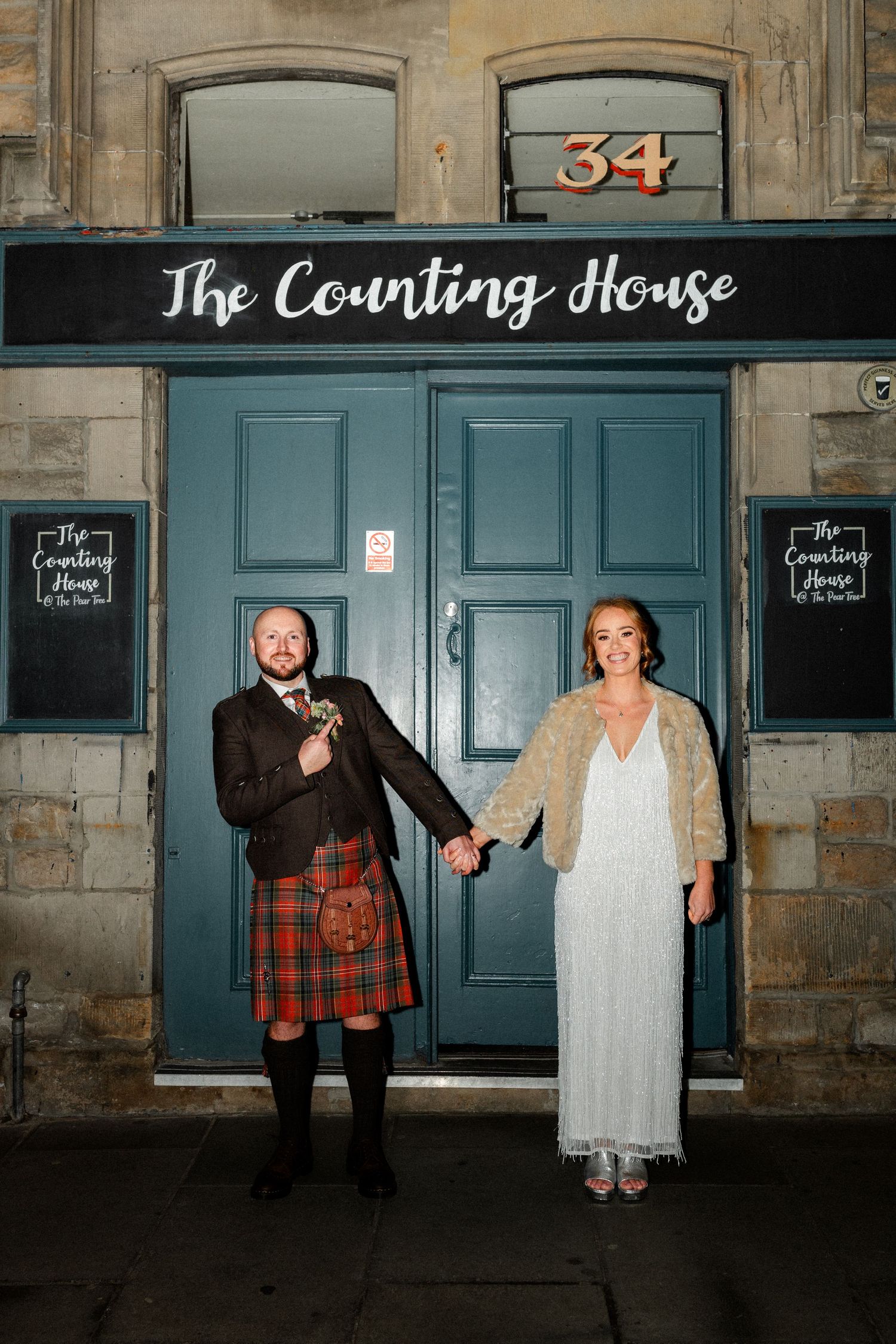 A bride and groom hold hands outside The Counting House venue, groom wearing a tartan kilt.