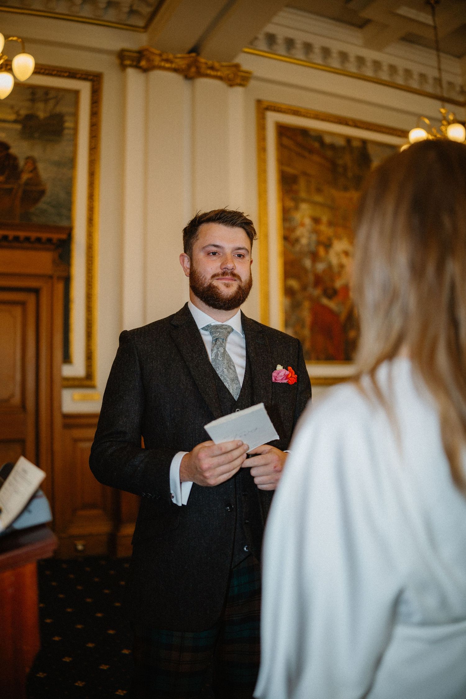 Groom in dark suit reading wedding vows to bride in an ornate gold and wood paneled ceremony room.