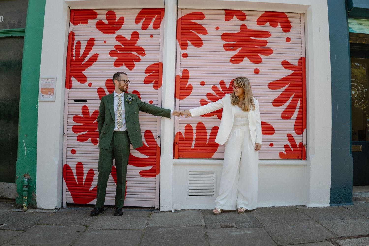 Bride and groom in stylish wedding attire posing playfully in front of a bold red floral painted shutter.