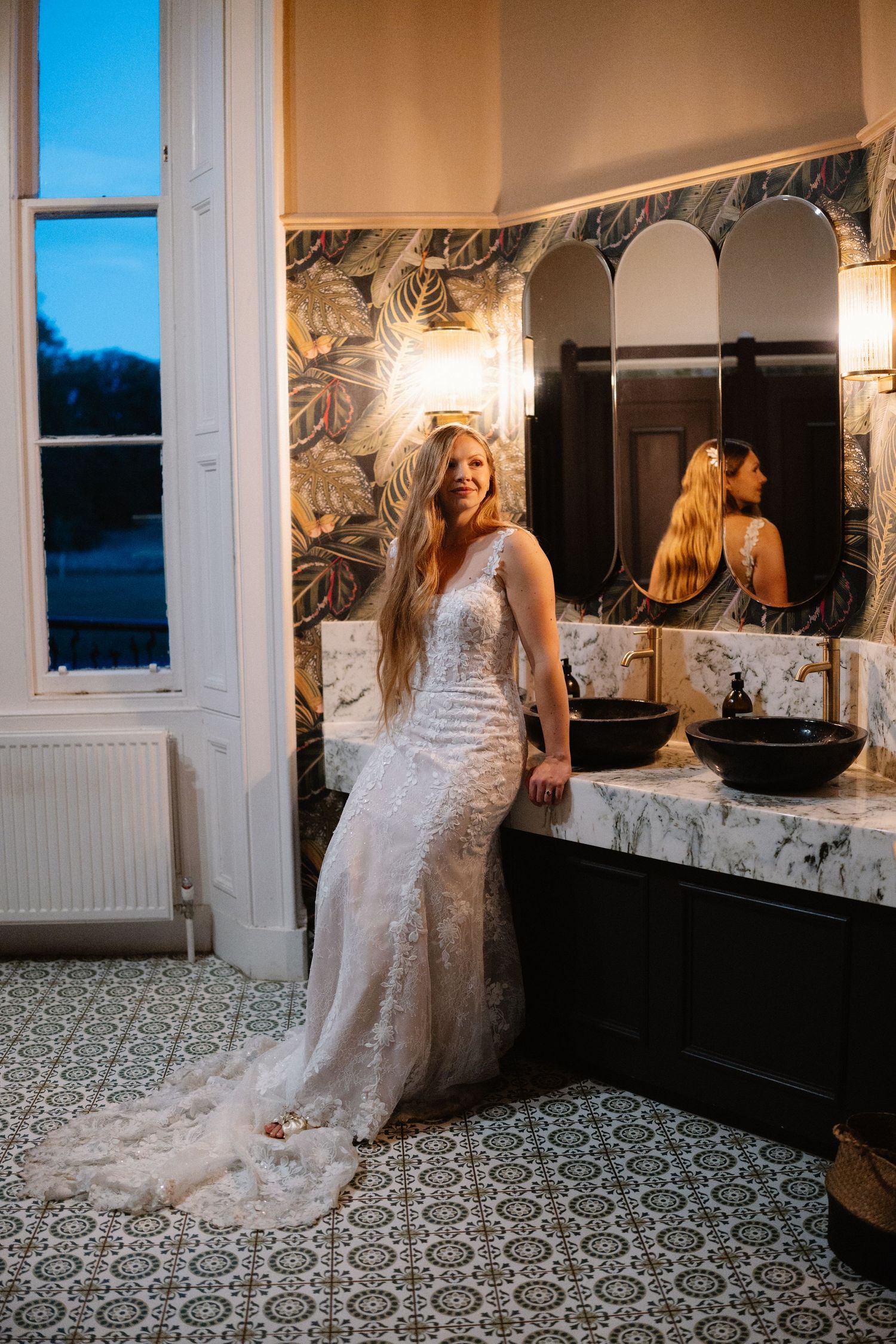 Bride in fitted lace wedding gown with long train posing in luxurious marble bathroom at evening wedding.