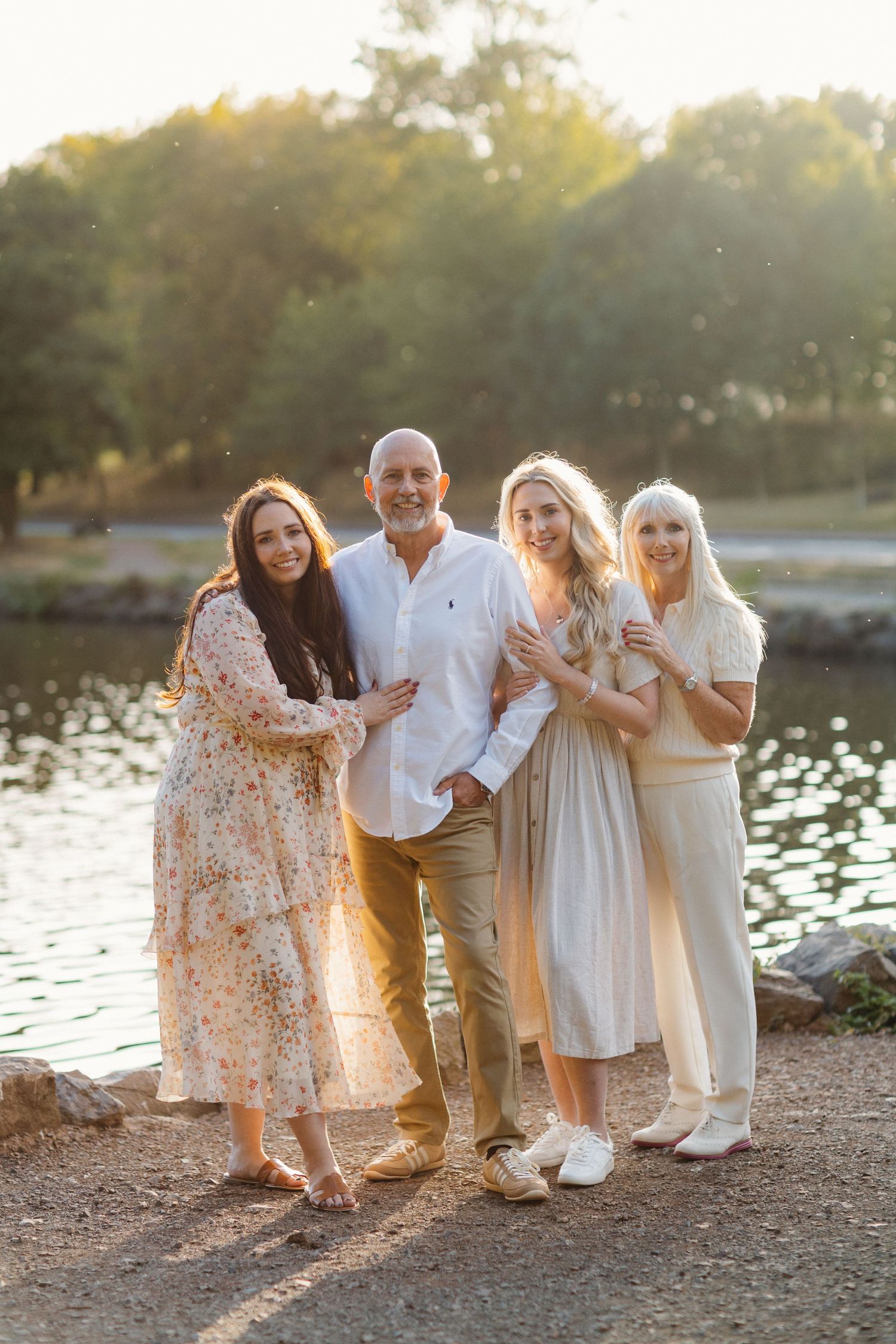 Family group wearing light colored clothing poses together by the water at sunset.