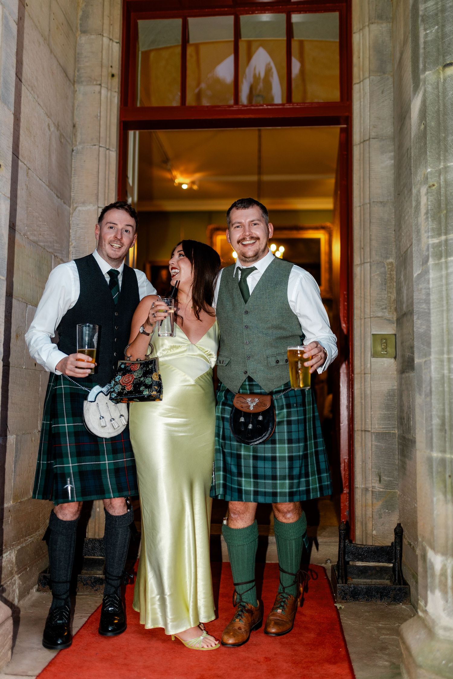 Wedding party members in traditional Scottish kilts and formal attire posing outside a stone building.