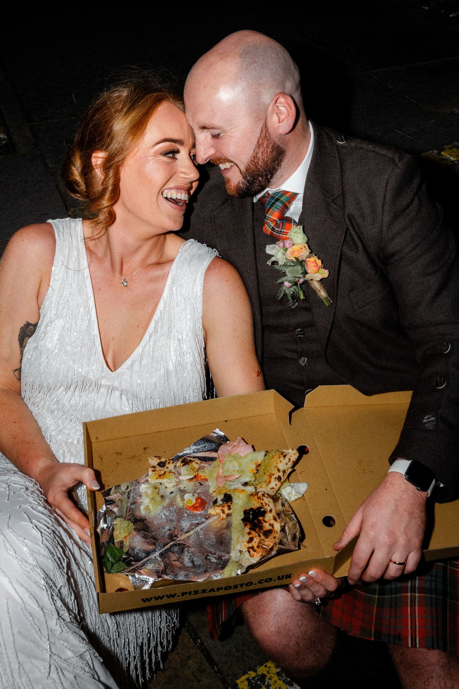 Couple in wedding attire sharing pizza from a cardboard box at night.