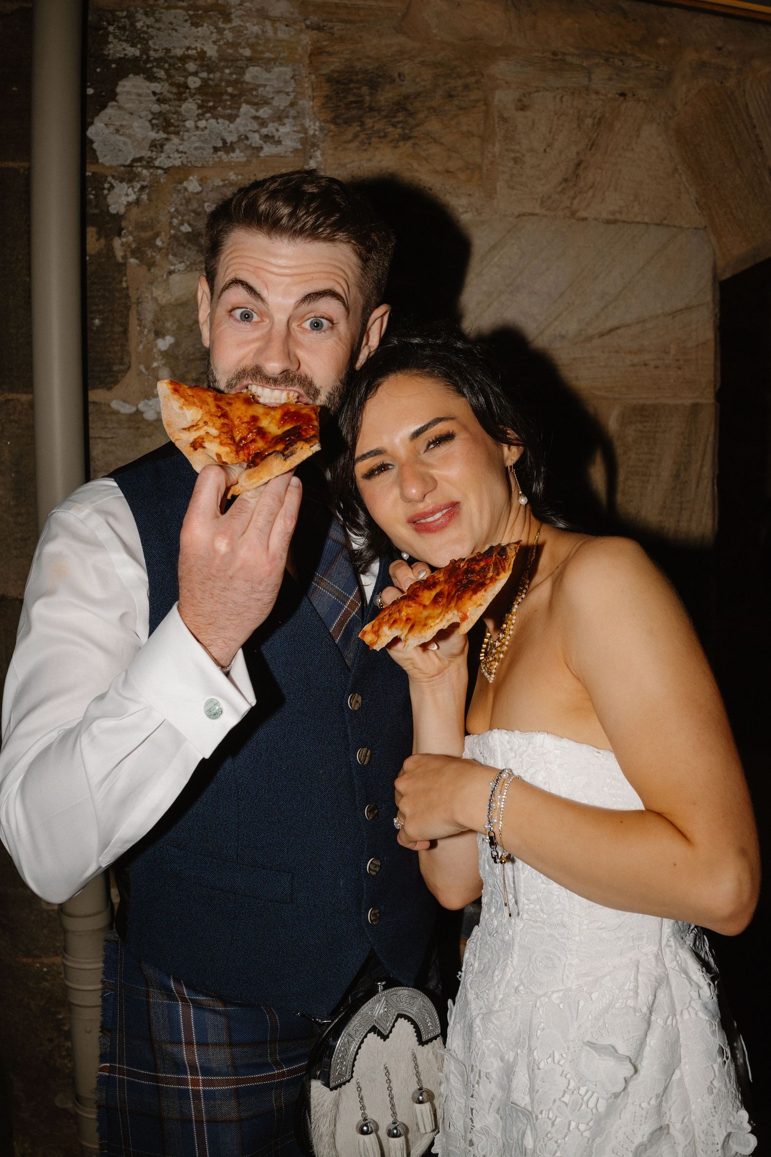Couple shares pizza and laughs during wedding reception in rustic venue.