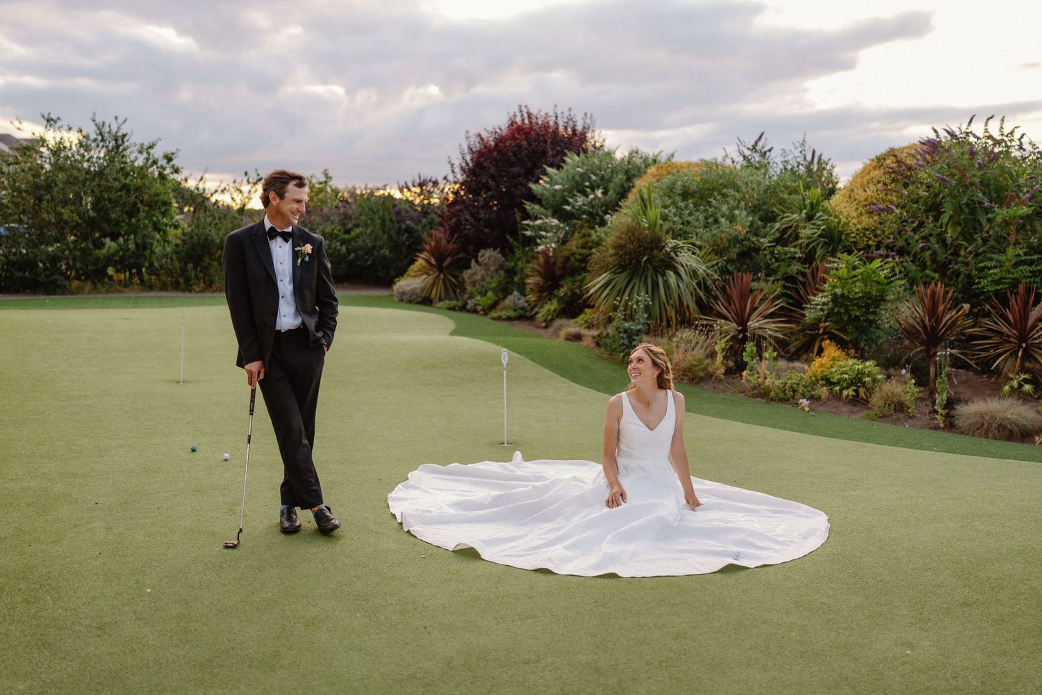 Bride in white gown sitting on golf green as groom in tuxedo putts at elegant garden wedding venue.
