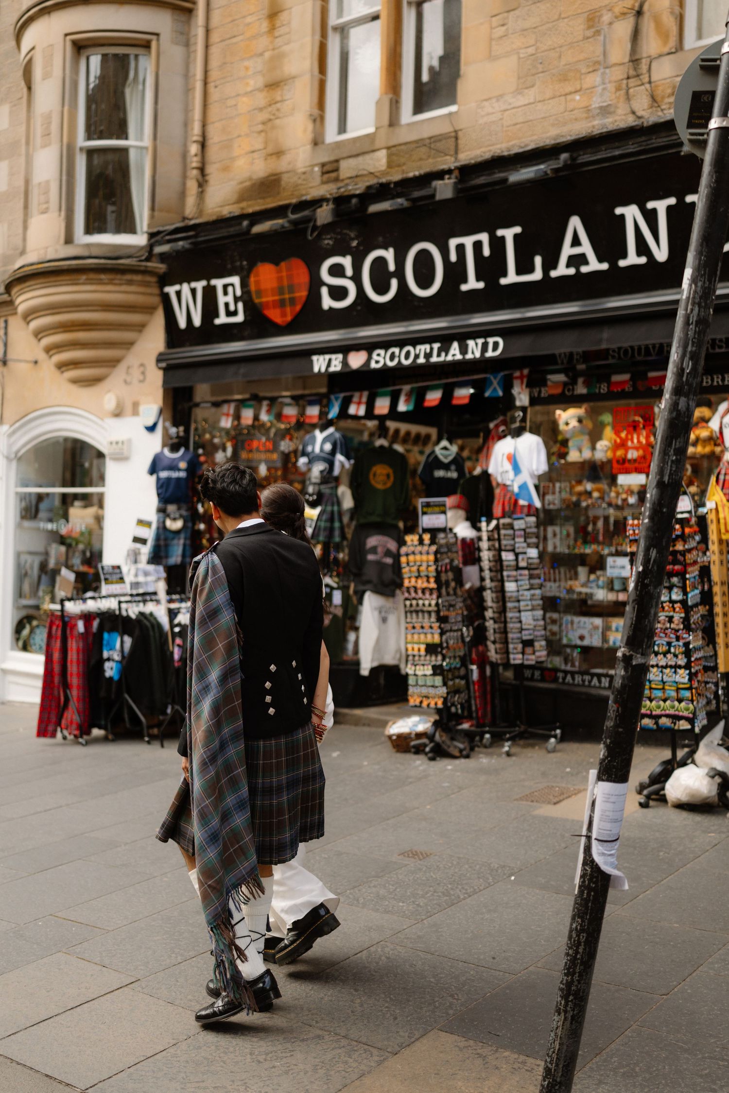 Groom in Scottish Highland kilt walking past a We Love Scotland souvenir shop on a city street.
